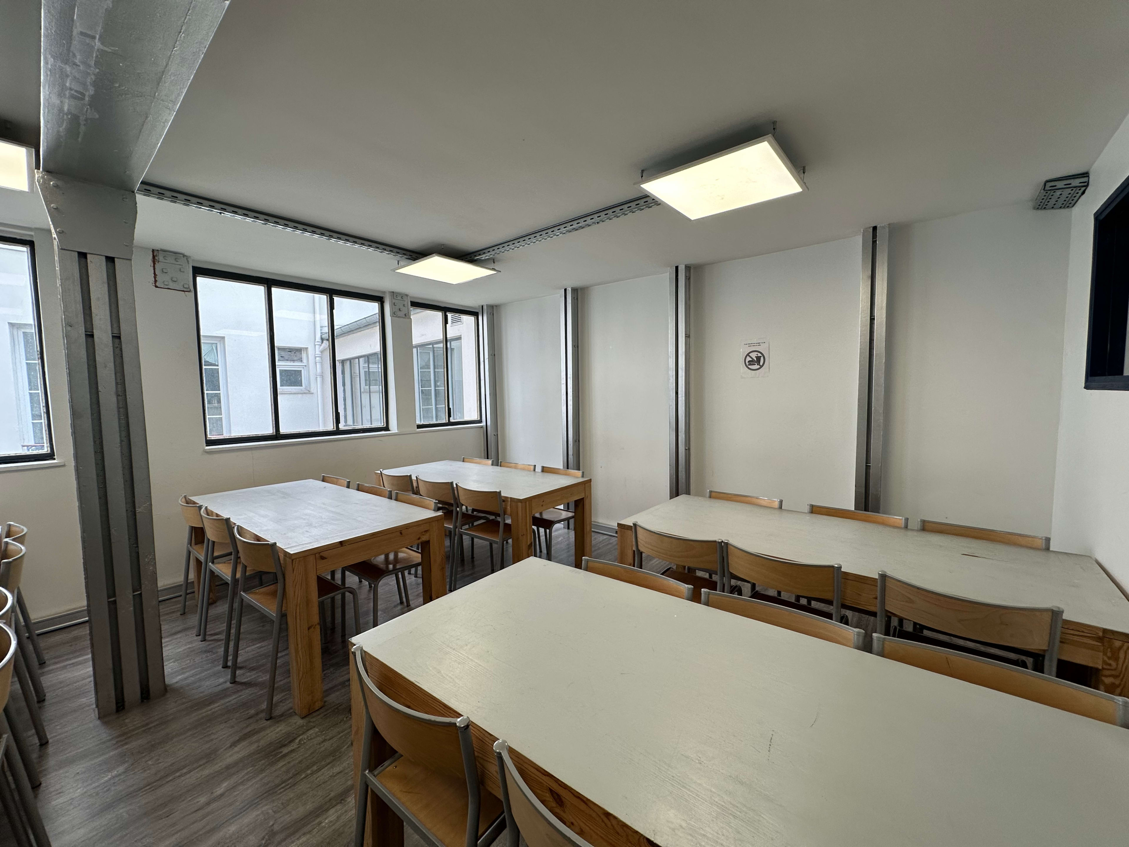 A room with multiple tables and chairs arranged in rows, illuminated by overhead lights and featuring large windows along the walls.