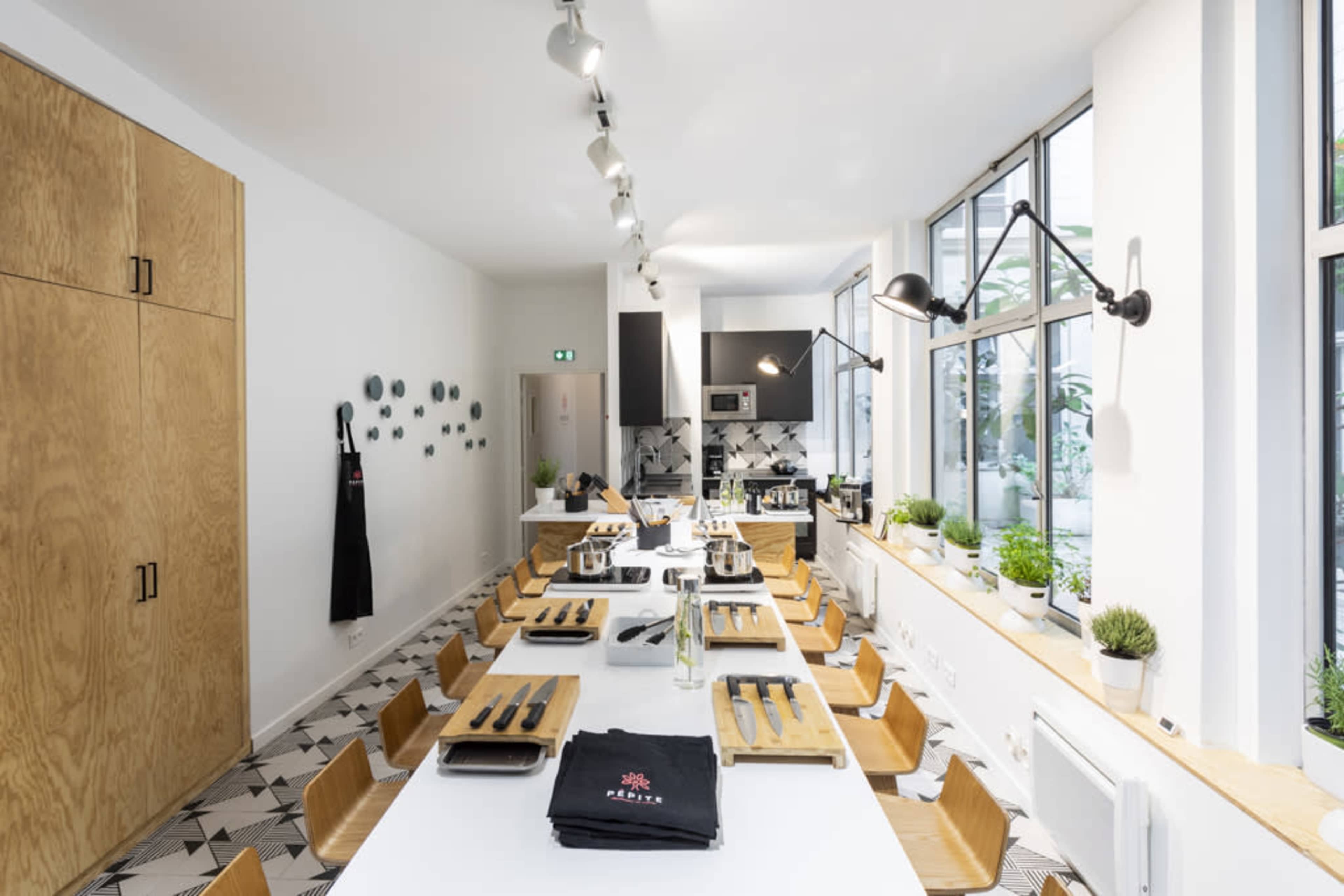 A modern kitchen with a long dining table set for a cooking class, featuring wooden chairs and various cooking utensils.