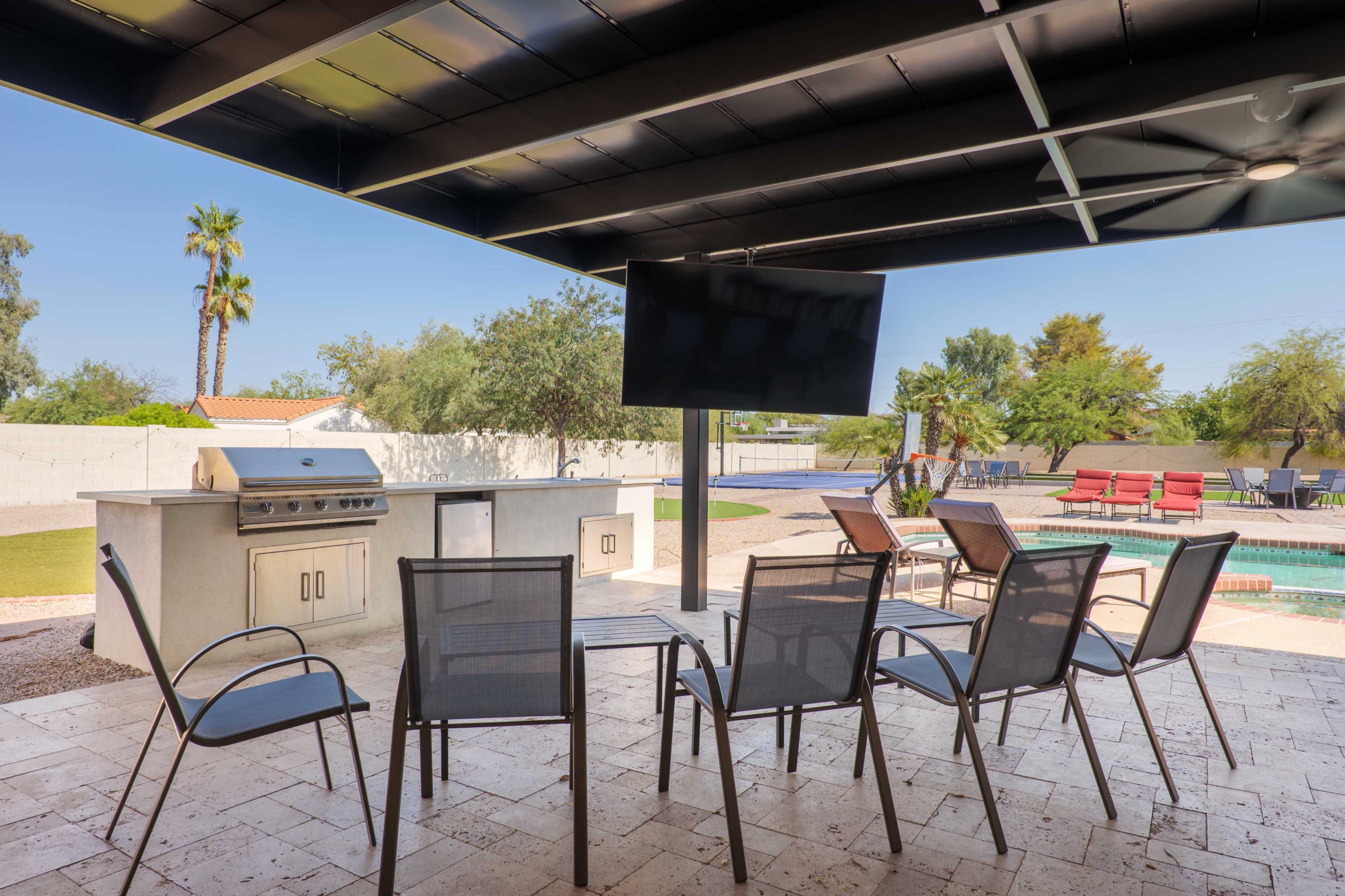 The image shows a covered outdoor patio with a barbecue grill, a large television mounted on the wall, and several chairs arranged around a patio table, overlooking a swimming pool and landscaped yard.