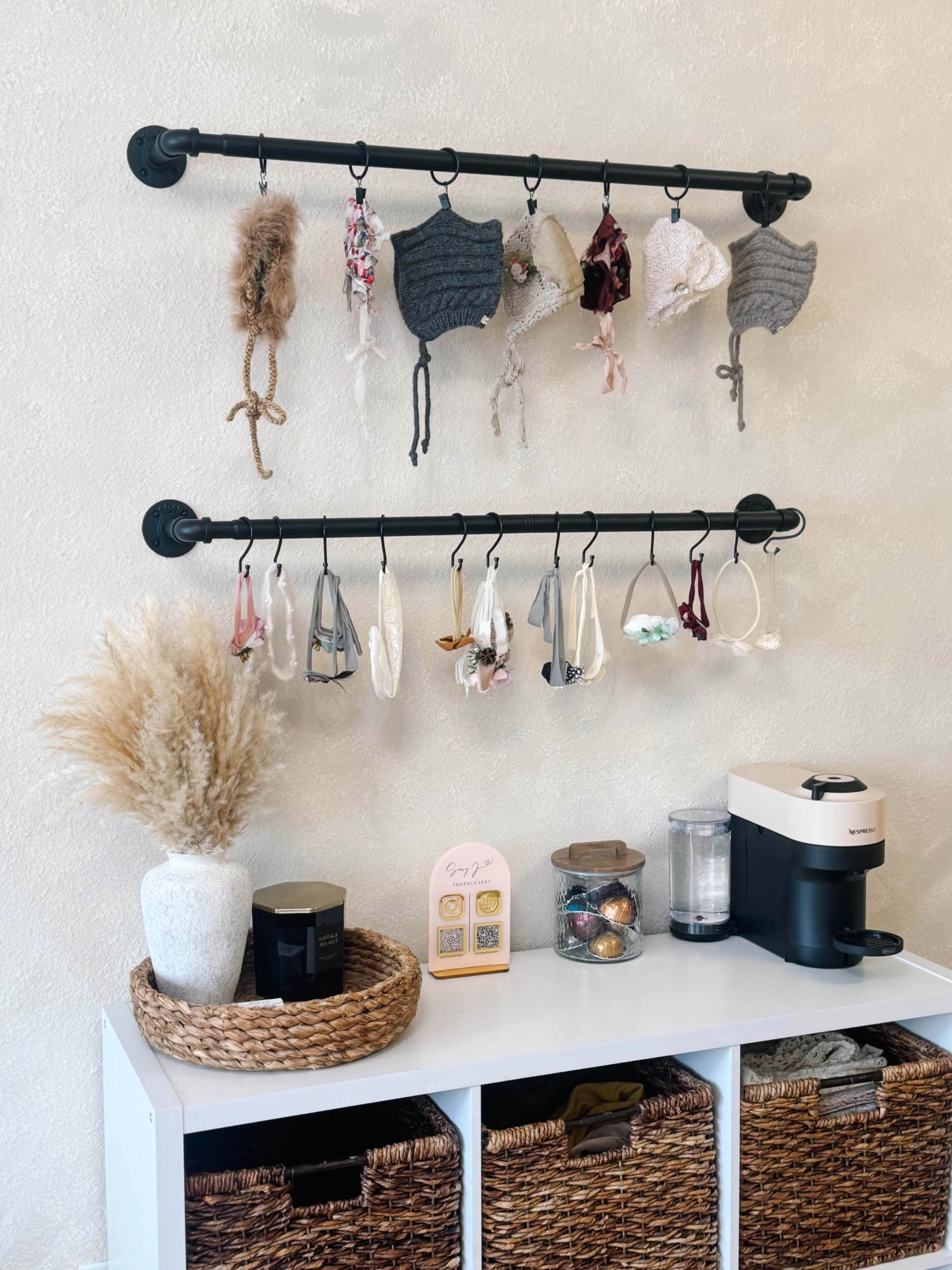 The image shows a wall-mounted display with two horizontal rods holding a variety of headbands and accessories, set above a countertop featuring decorative items, a coffee maker, and storage baskets.