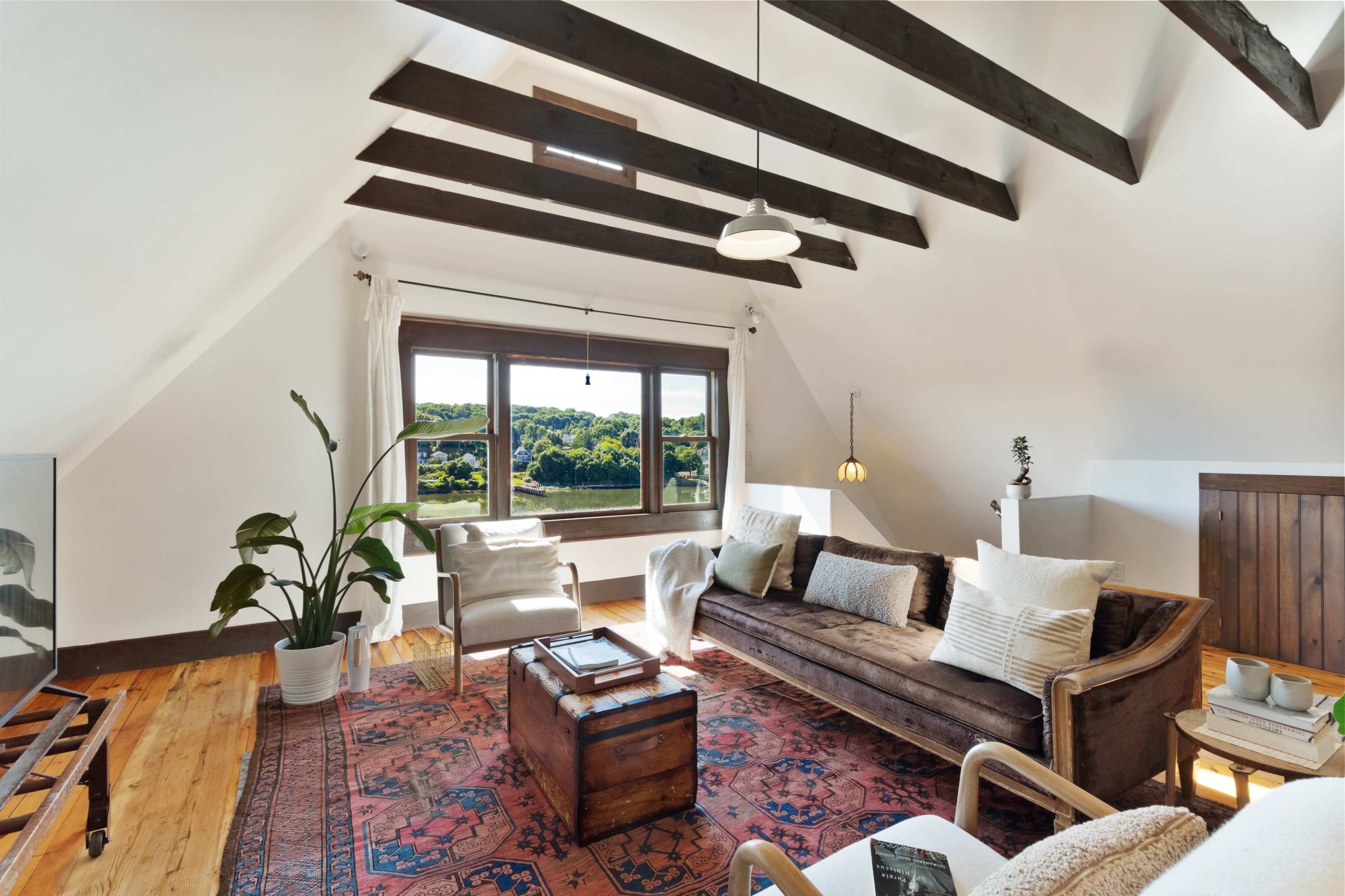The image shows a cozy living room with a brown sofa, an area rug, a wooden coffee table, and large windows that overlook a green landscape.