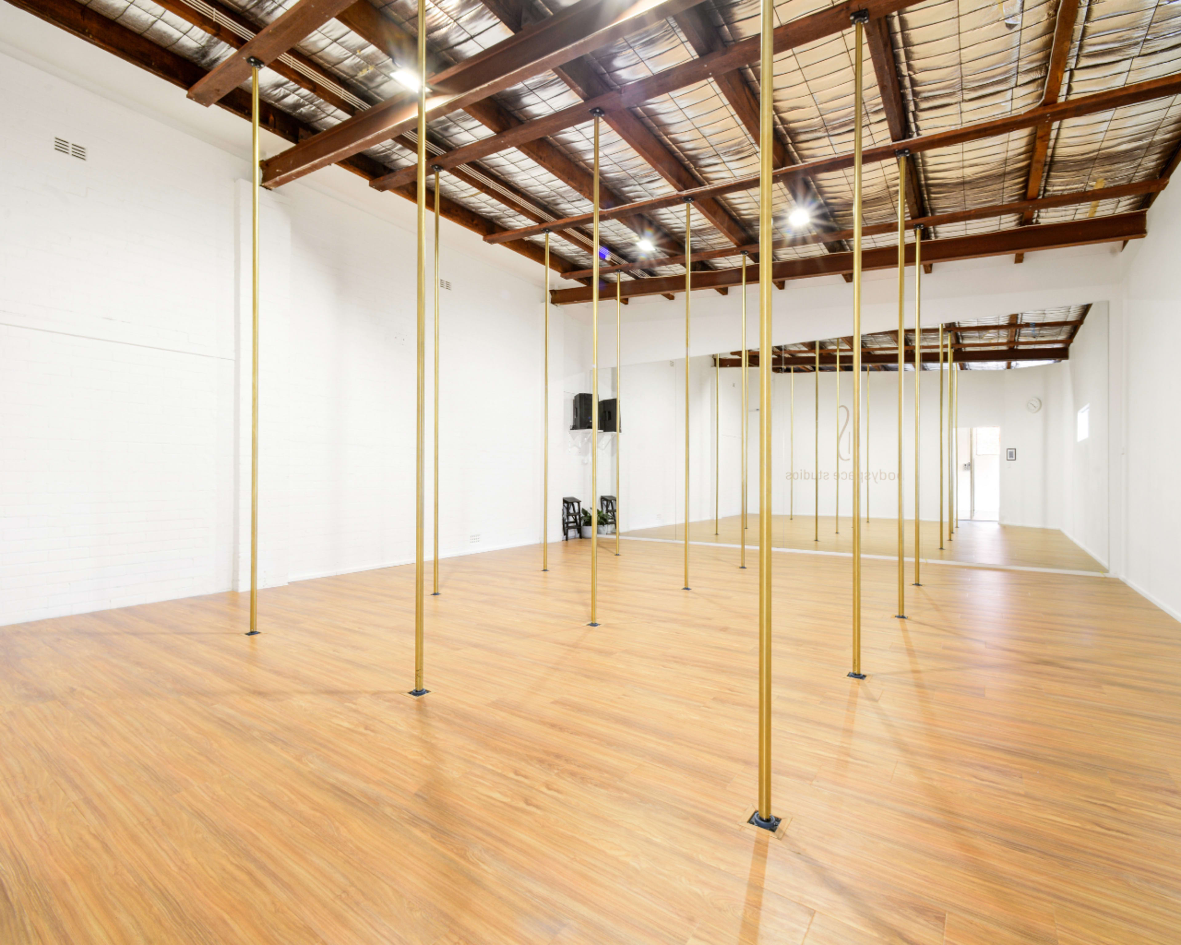 The image shows an empty dance studio with wooden flooring and multiple gold poles spaced evenly throughout the room.