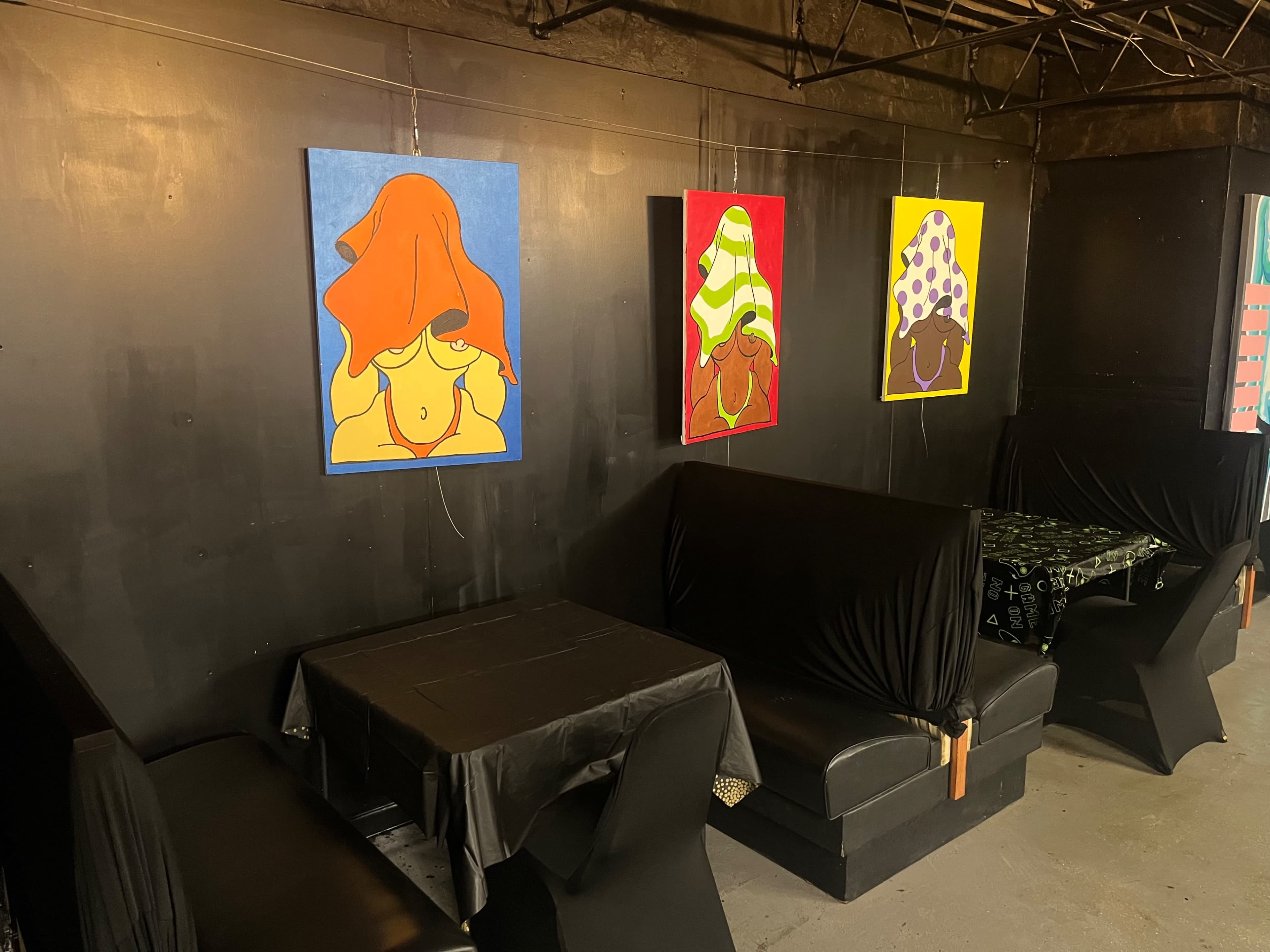A dark-walled room with four colorful paintings of women with head coverings displayed on the wall above two black-covered tables and seating areas.