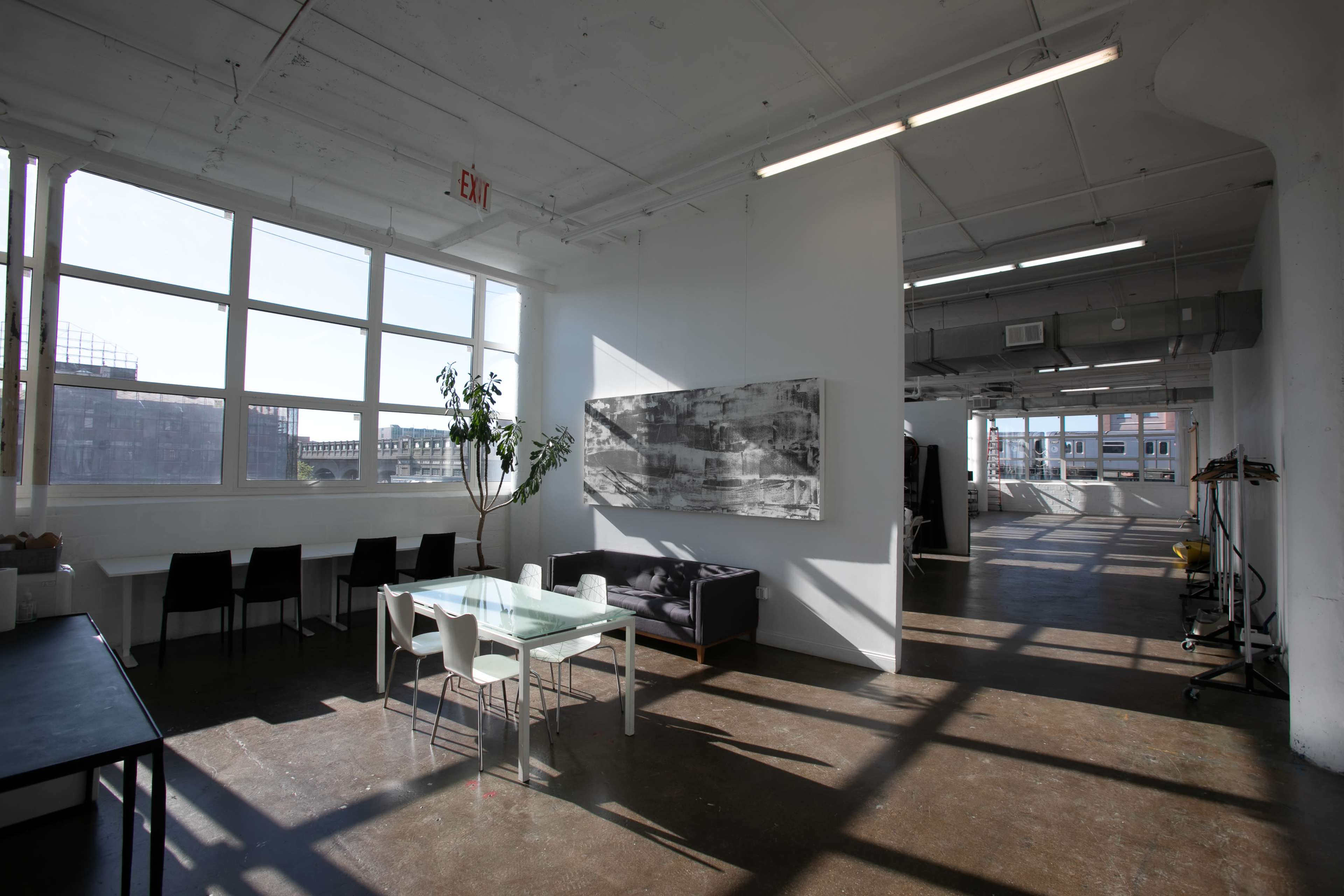 A modern, open office space features large windows, a dining table, chairs, and artwork on the wall.