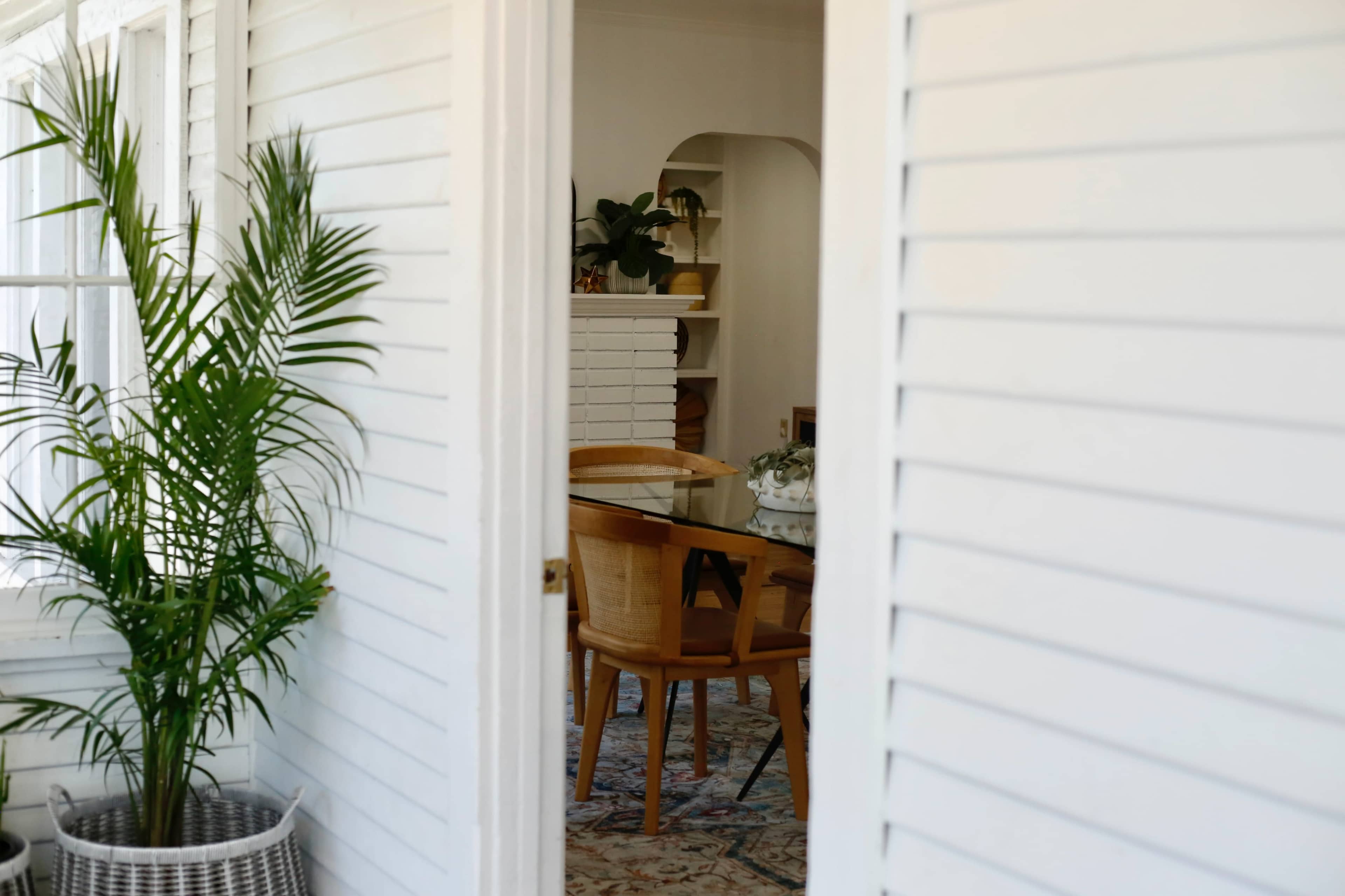 A glimpse of a bright interior space with a glass dining table and wooden chairs, framed by white wooden shutters and a potted plant in the foreground.