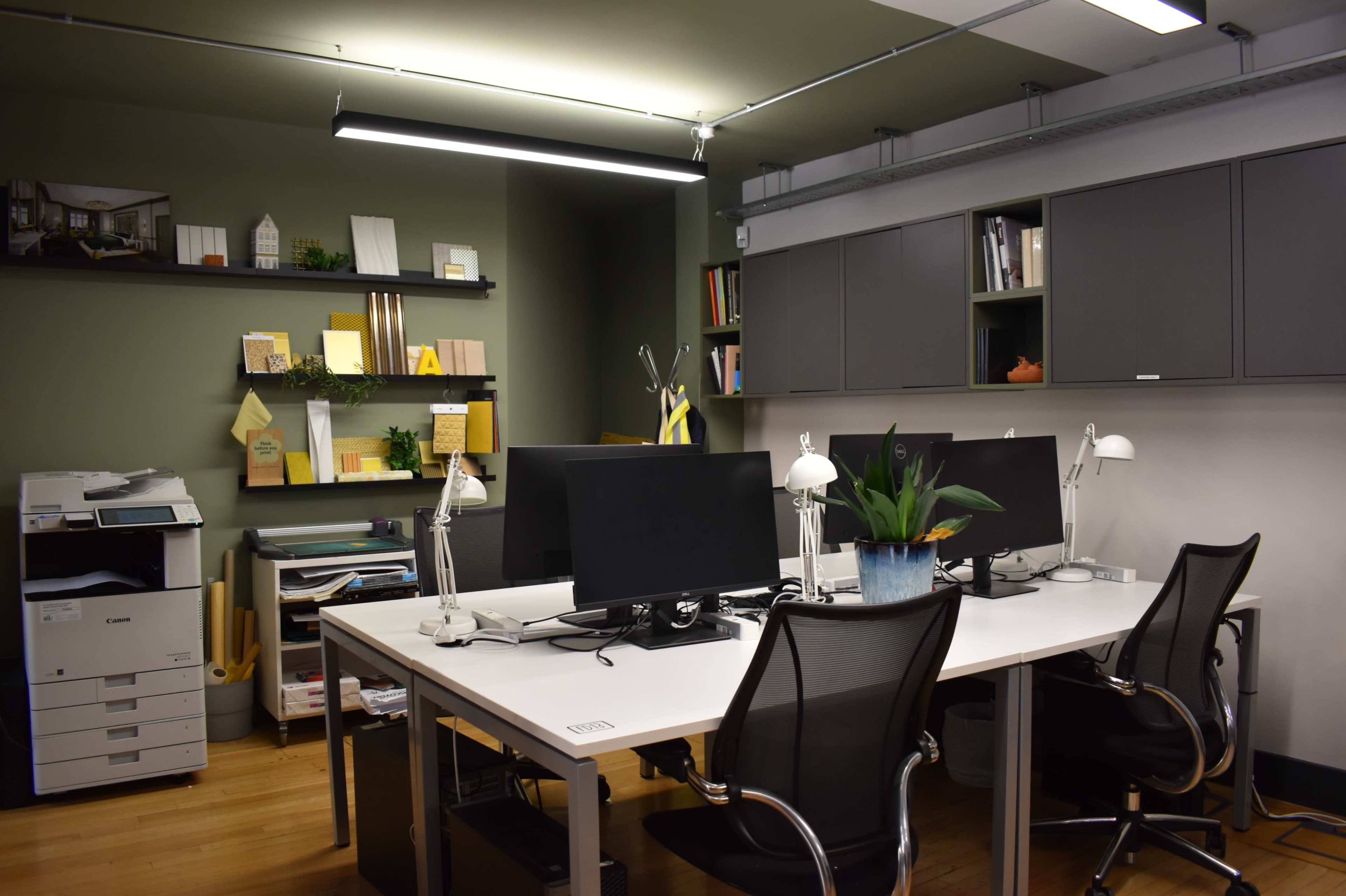 The image shows a modern office space featuring two workstations with monitors, an indoor plant, and shelves filled with books and materials.