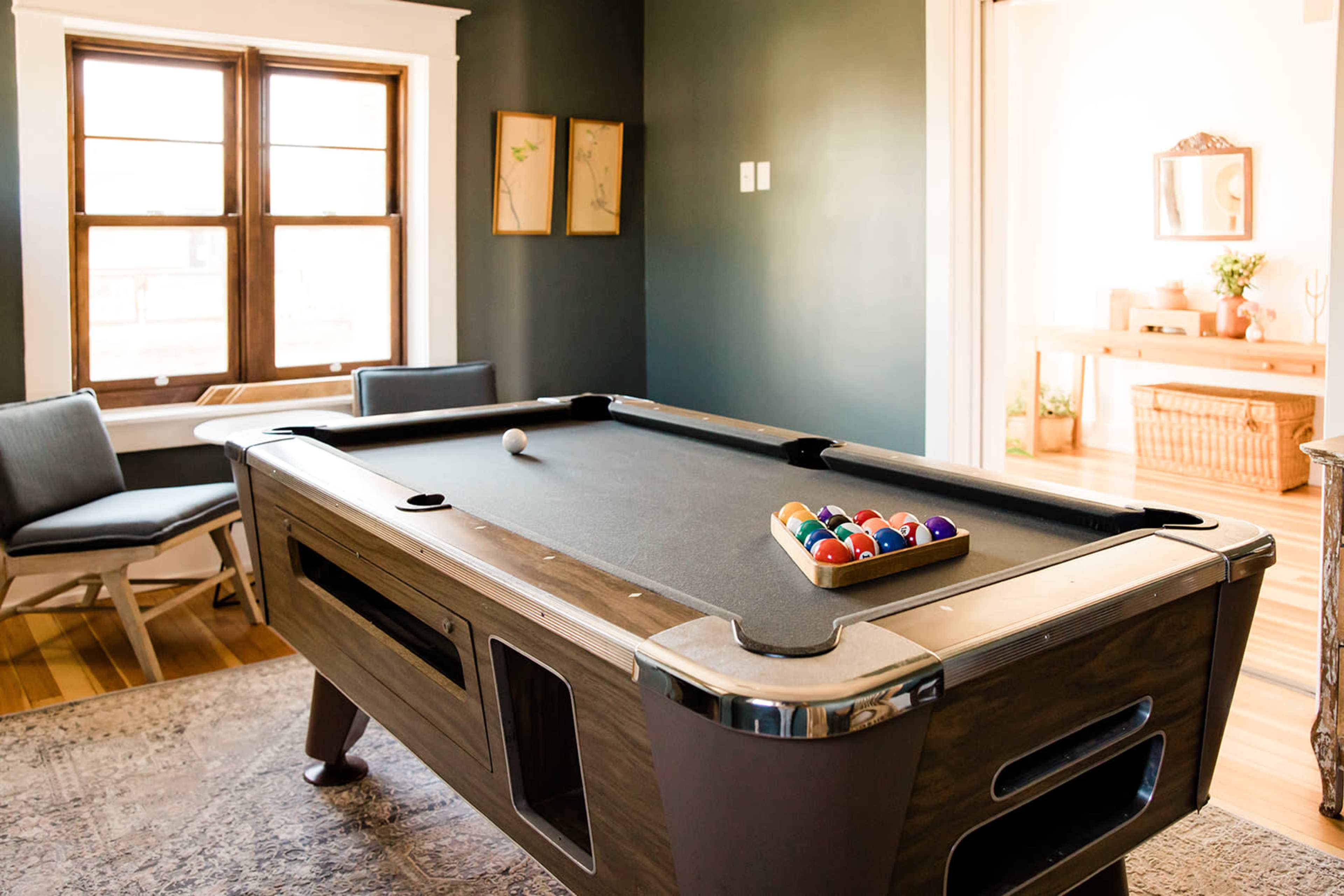 A pool table with a full rack of balls is positioned in a room featuring green walls and large windows.