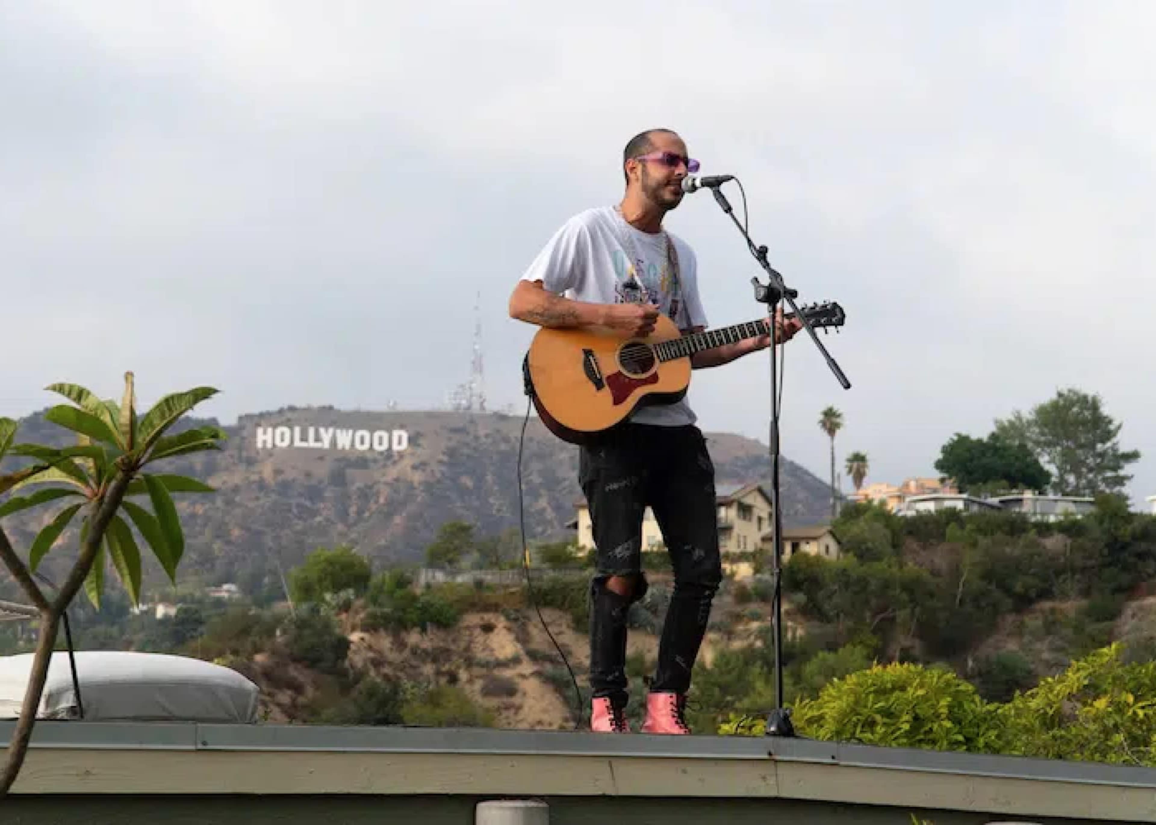A musician plays guitar and sings on a rooftop with the Hollywood sign in the background.