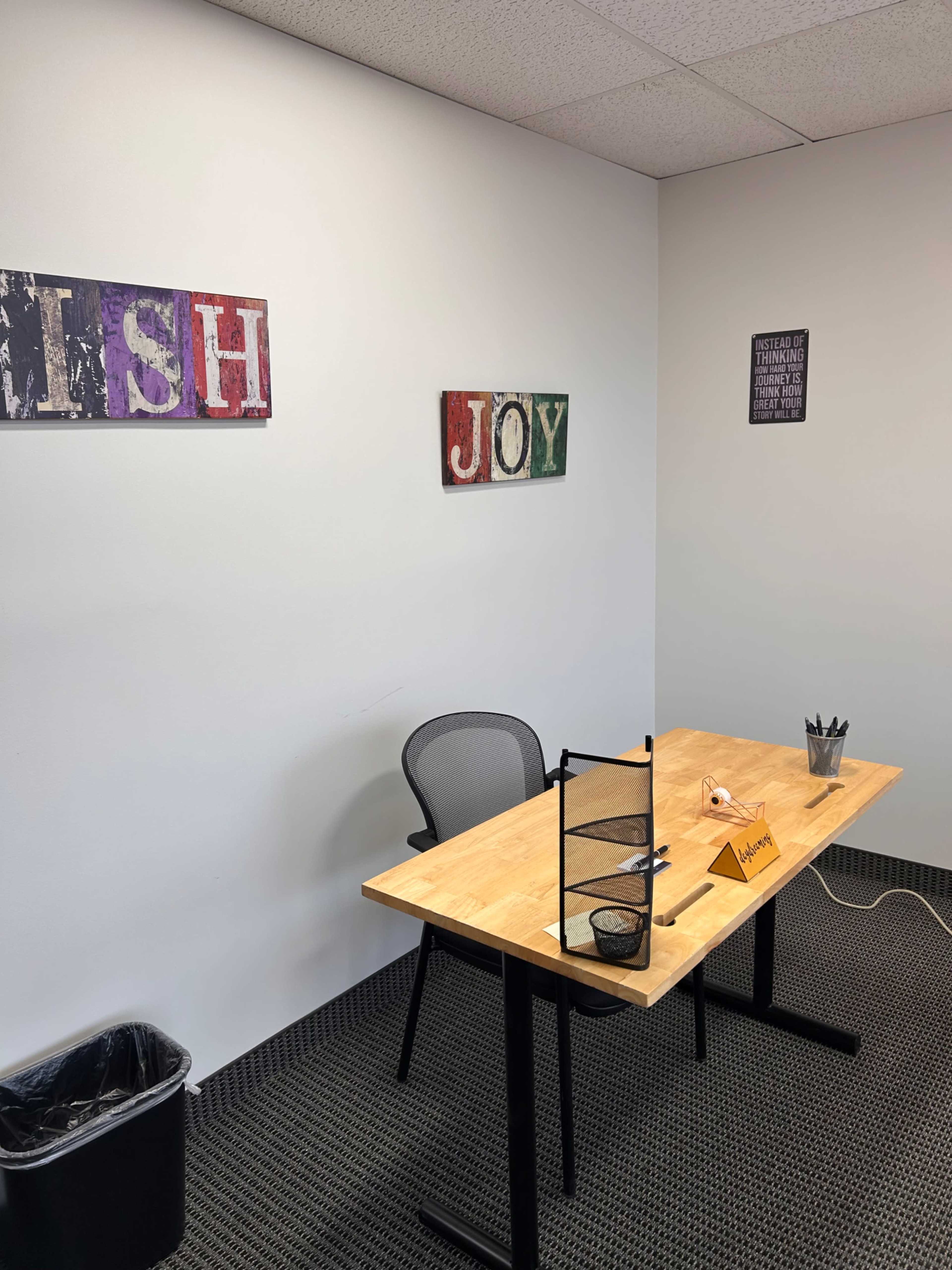 The image shows a small, empty office space with a wooden desk, a black chair, a trash can, and framed signs on the wall displaying the words "WISH" and "JOY."