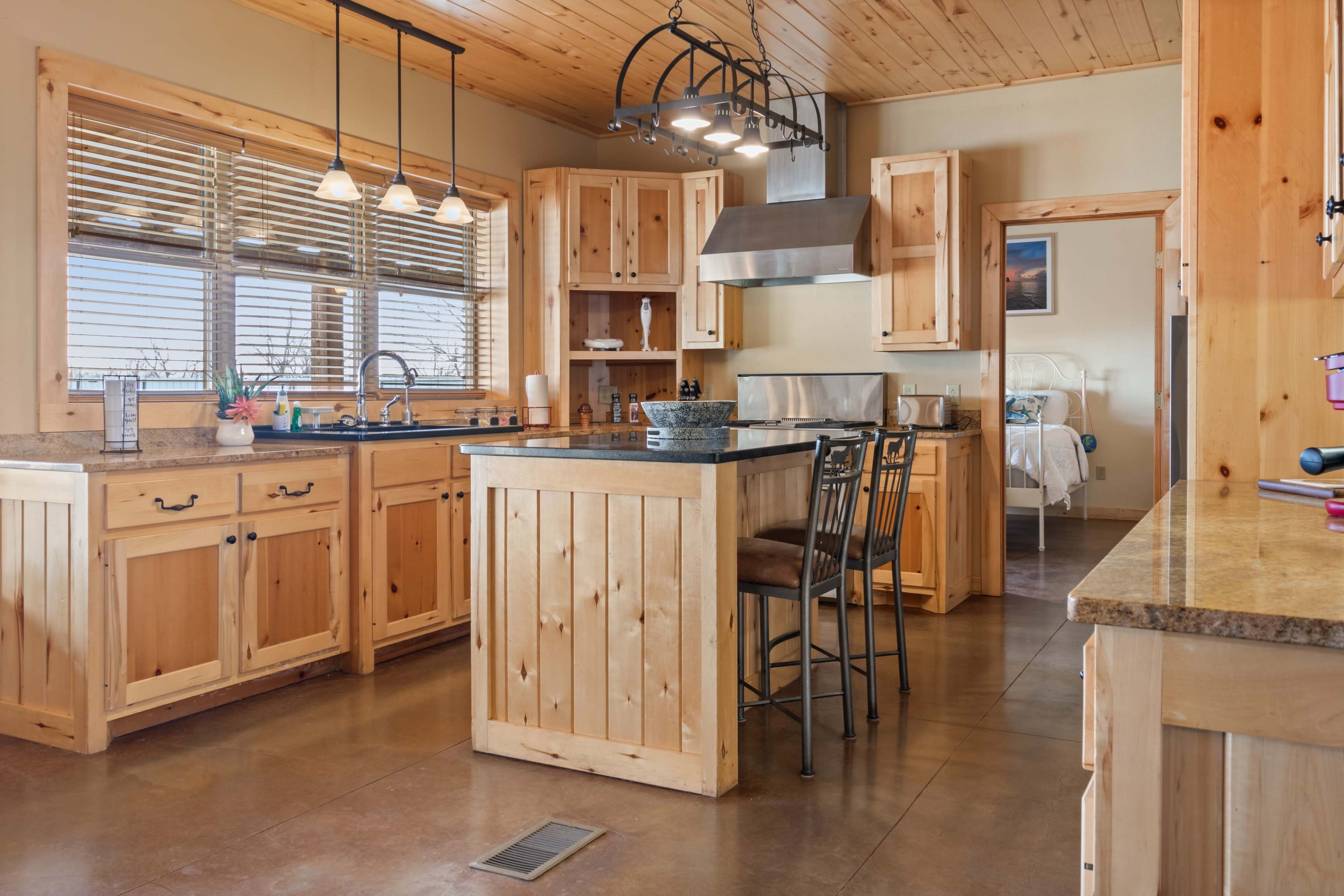 A wooden kitchen with a central island, bar stools, and modern appliances, featuring a warm color scheme and a view of a bedroom through an open door.