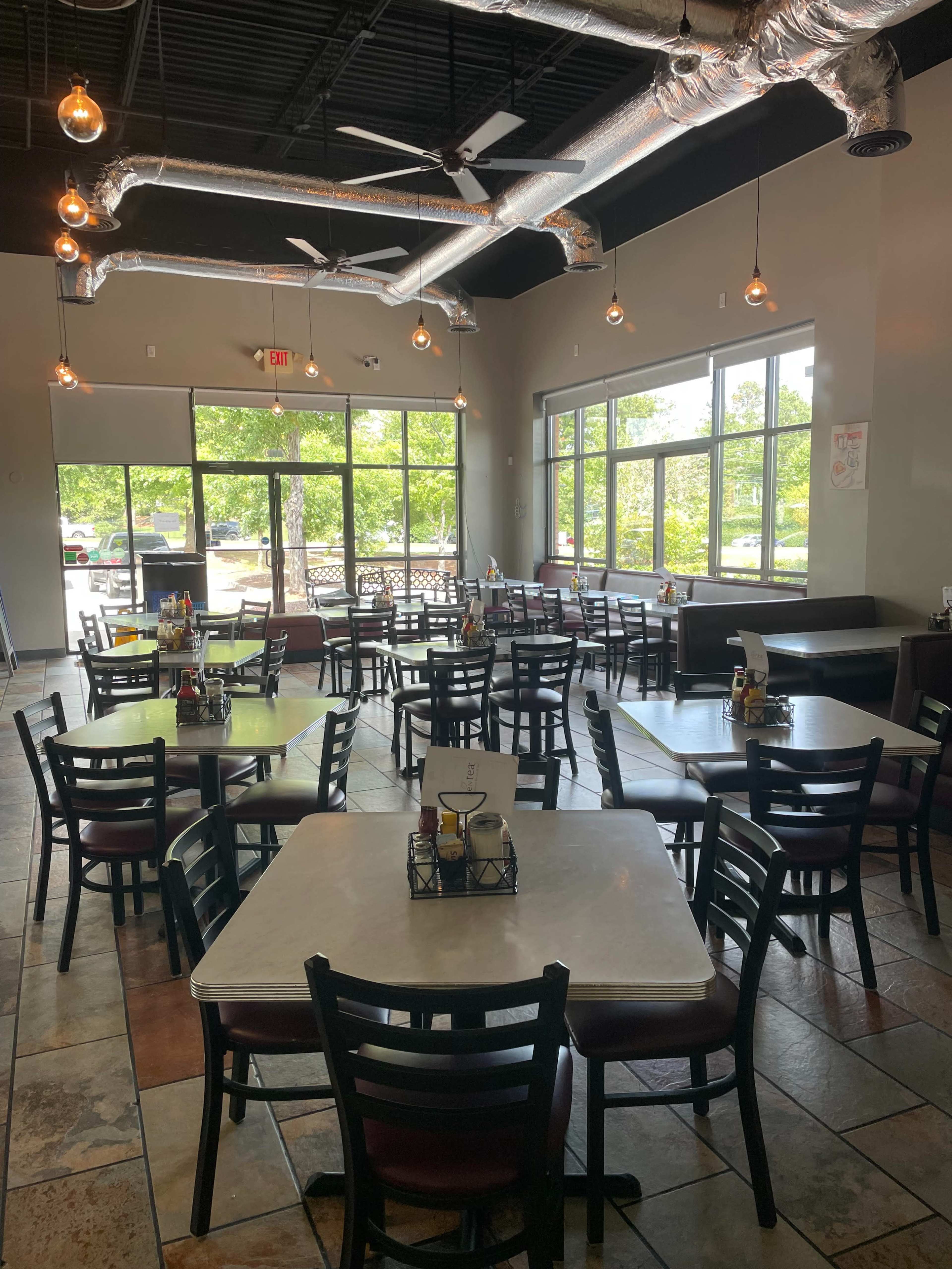 The image shows an empty restaurant interior with multiple tables and chairs arranged around a central area, large windows letting in natural light, and industrial-style lighting fixtures.