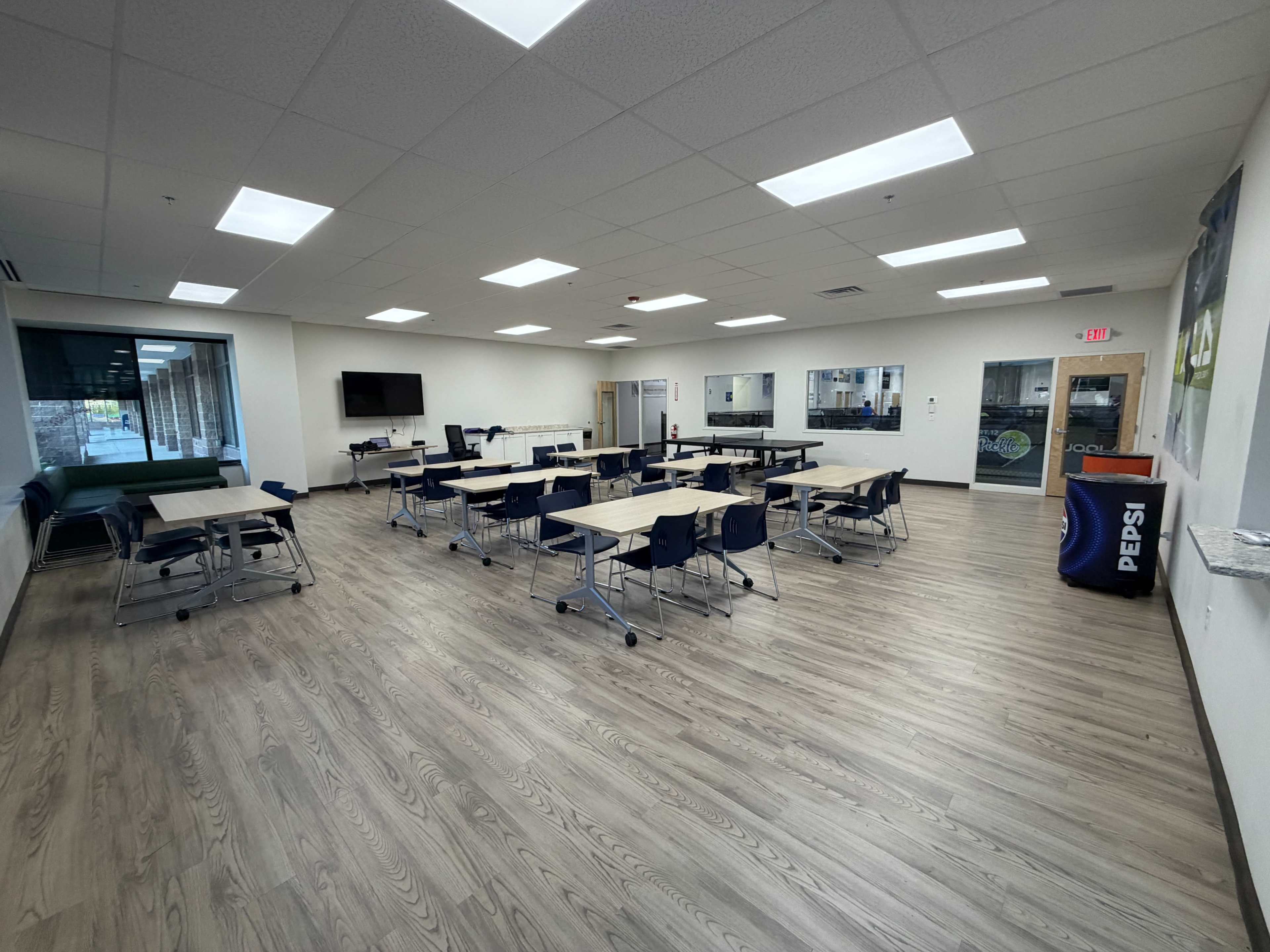 The image shows a spacious classroom with multiple tables and chairs arranged for group work, along with a television on the wall and a beverage station in the corner.