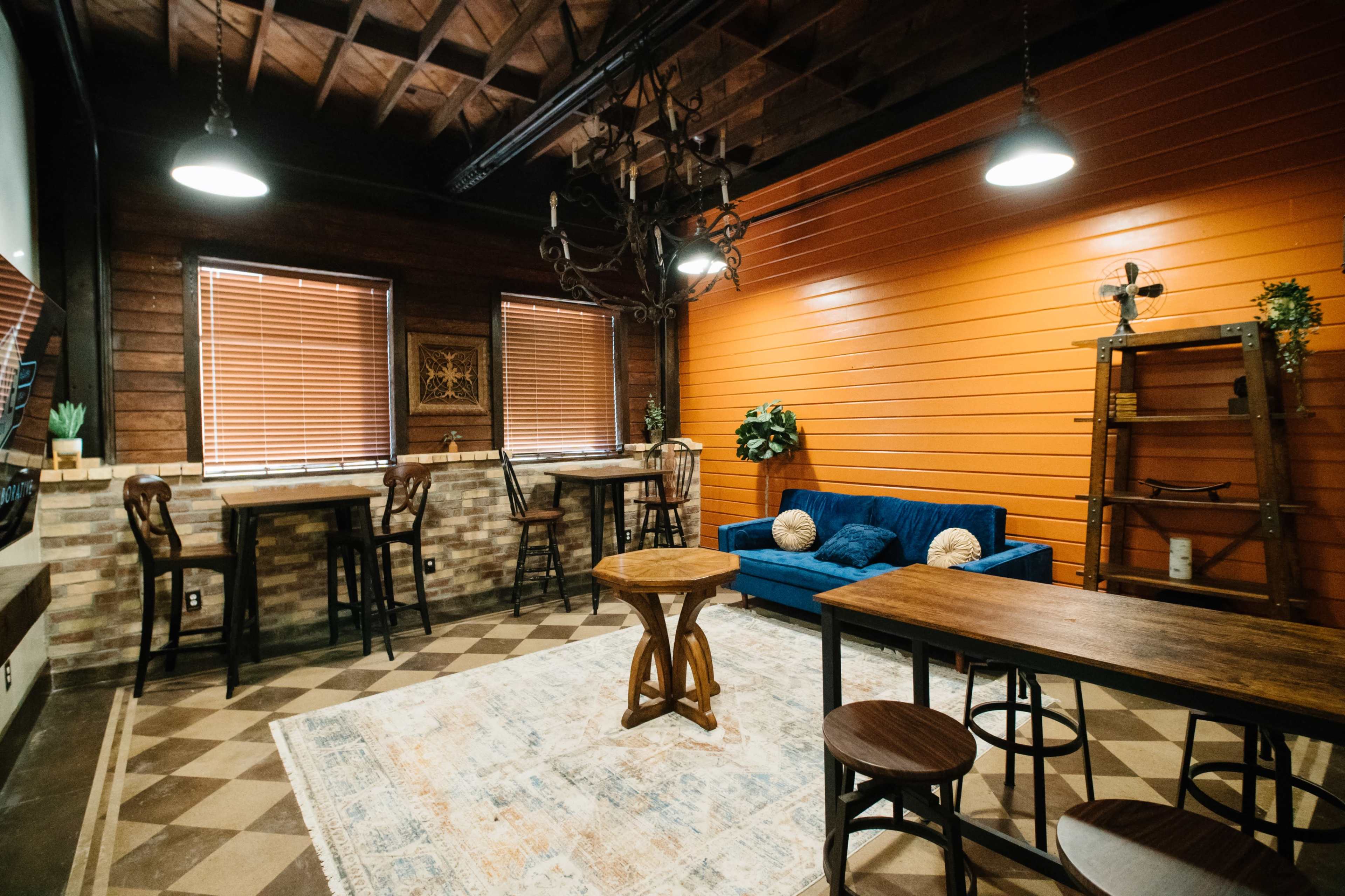 A cozy room featuring a blue couch, several wooden tables and chairs, a patterned rug, and orange accent walls with window blinds.