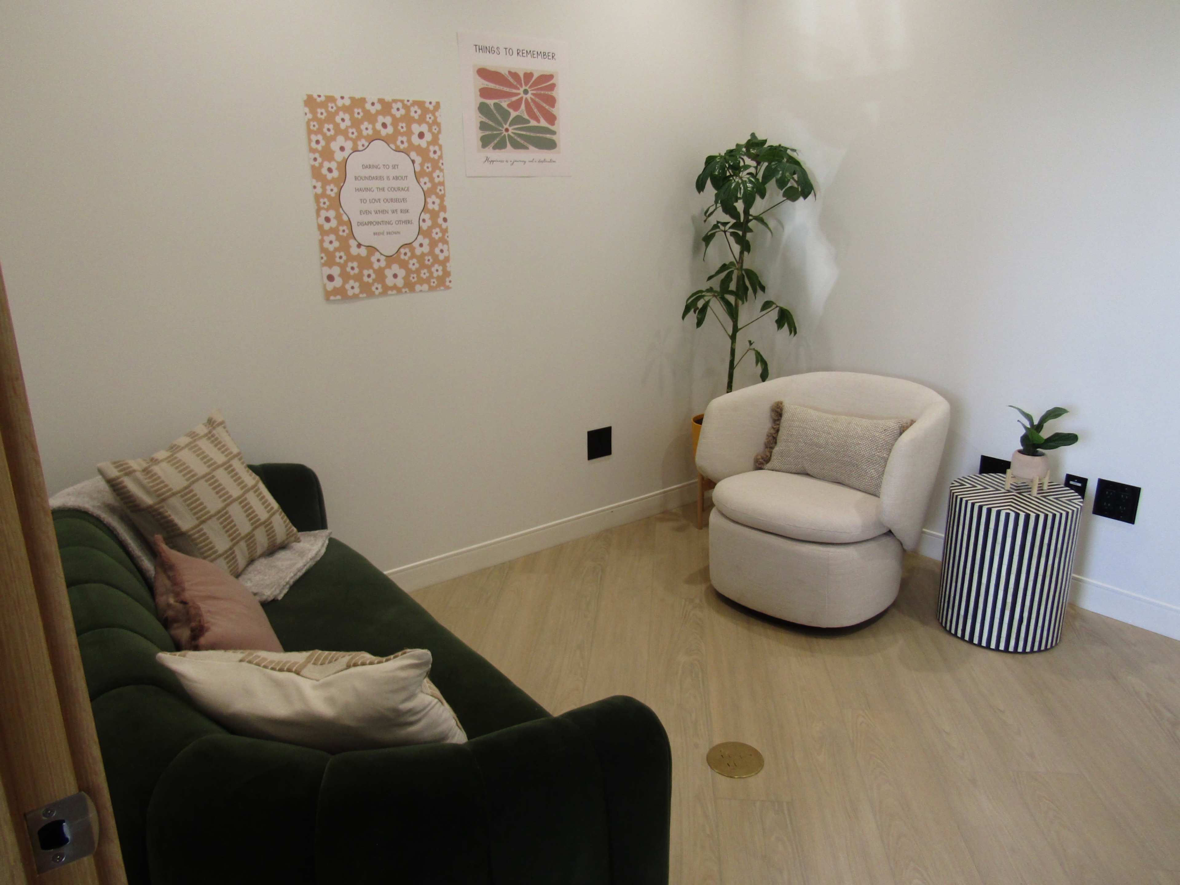 The image shows a small, well-lit room featuring a green sofa, a white armchair, a small striped table, and a plant, with decorative posters on the walls.