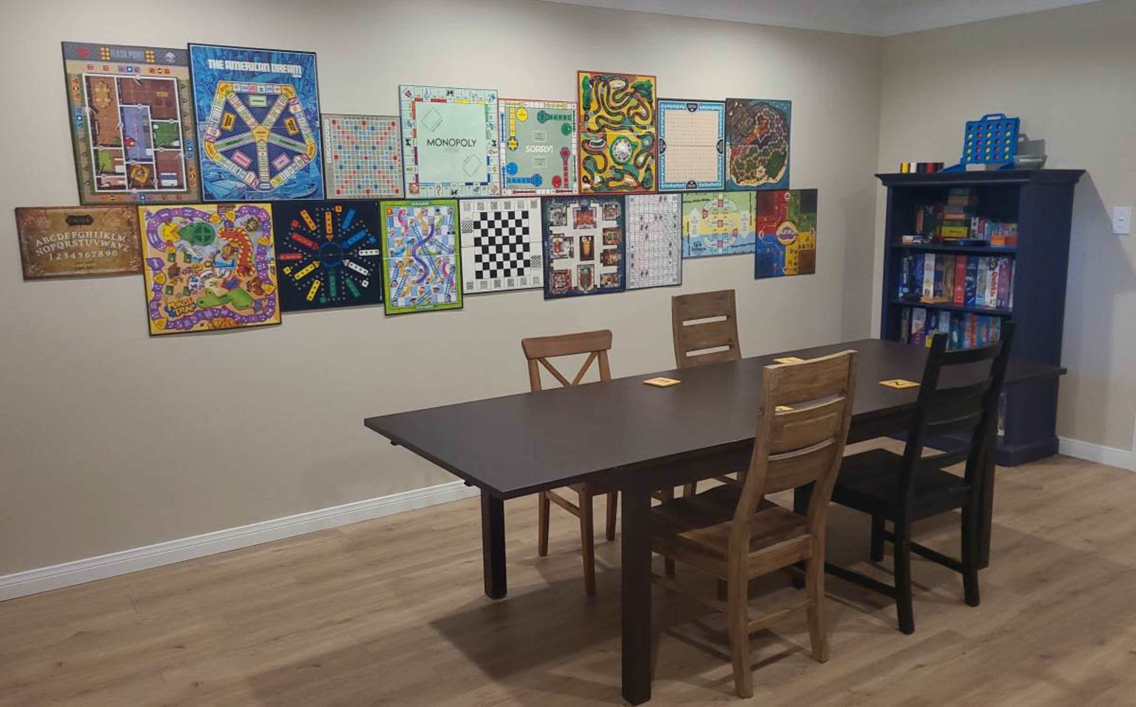 A game room features a large table surrounded by chairs, with various board games displayed on the wall and a bookshelf filled with games in the corner.