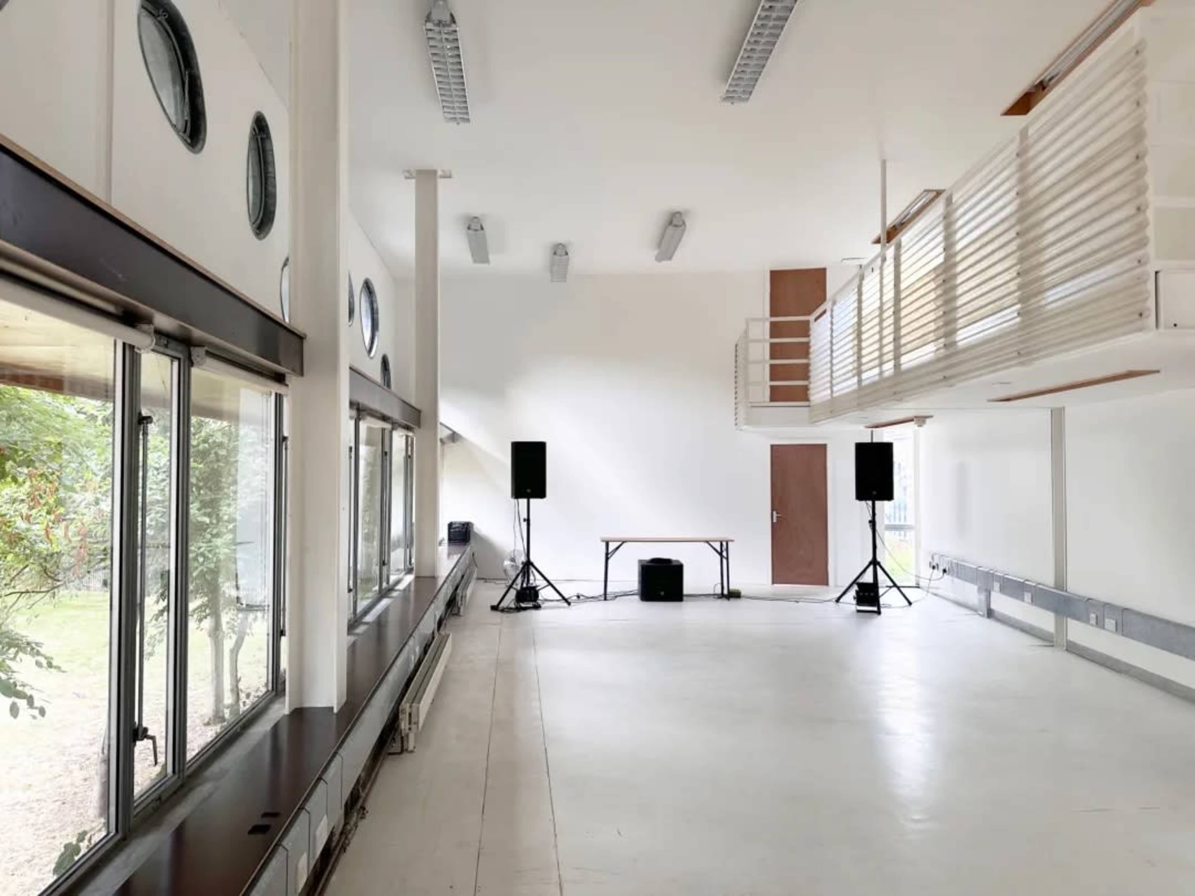 The image shows a spacious, well-lit room with large windows, featuring a balcony, audio equipment, and minimal furnishings.