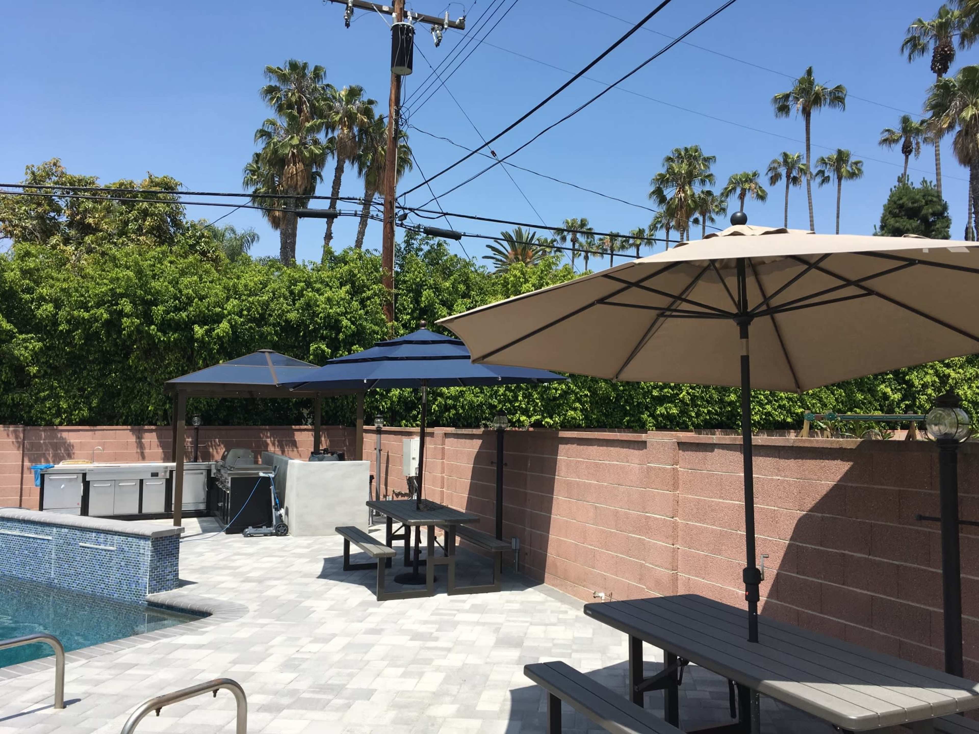 A patio area with two large umbrellas, a rectangular pool, several tables, and palm trees in the background under a clear blue sky.