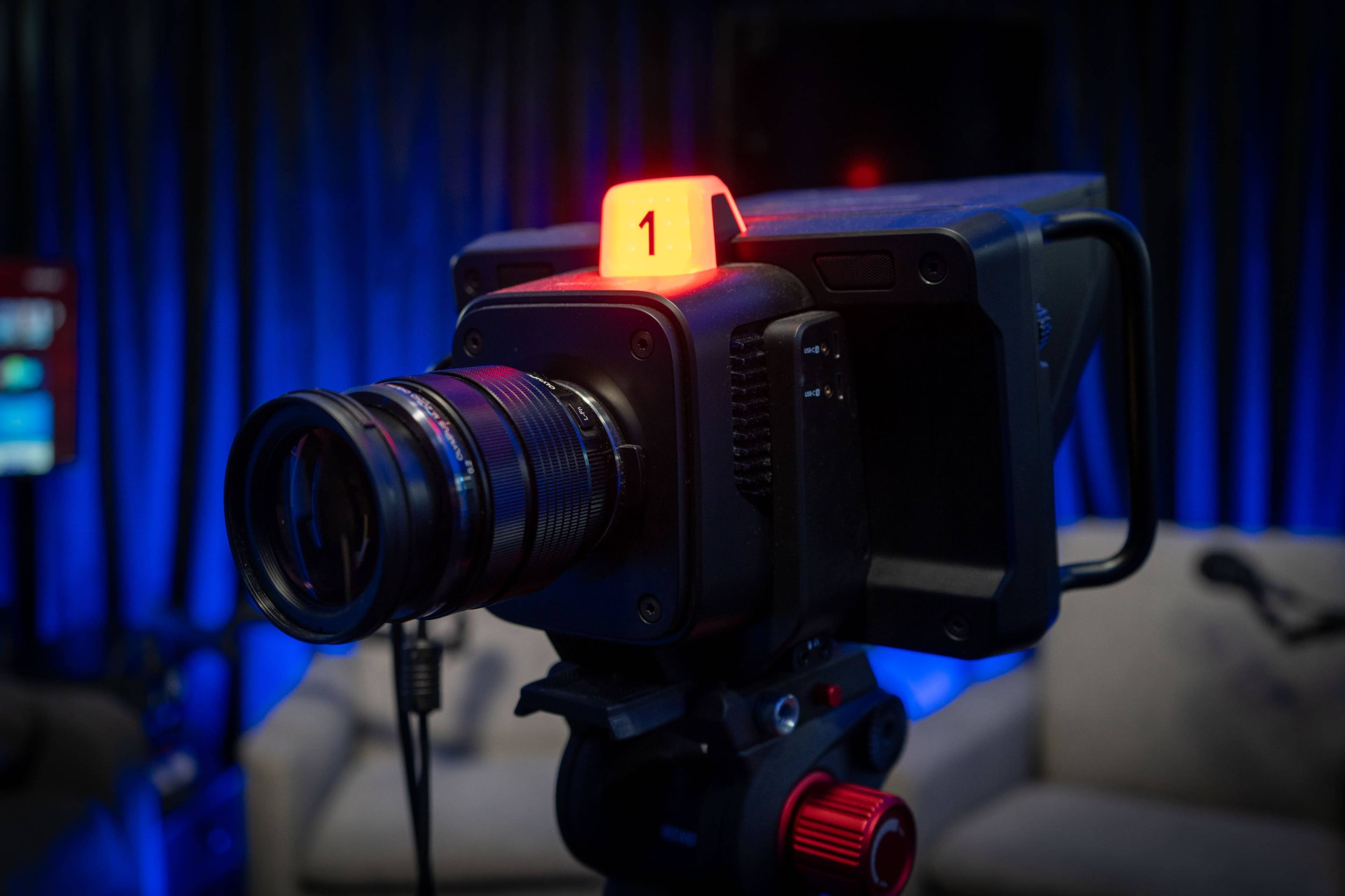 A professional camera with a red indicator light sits on a tripod in a studio setting.