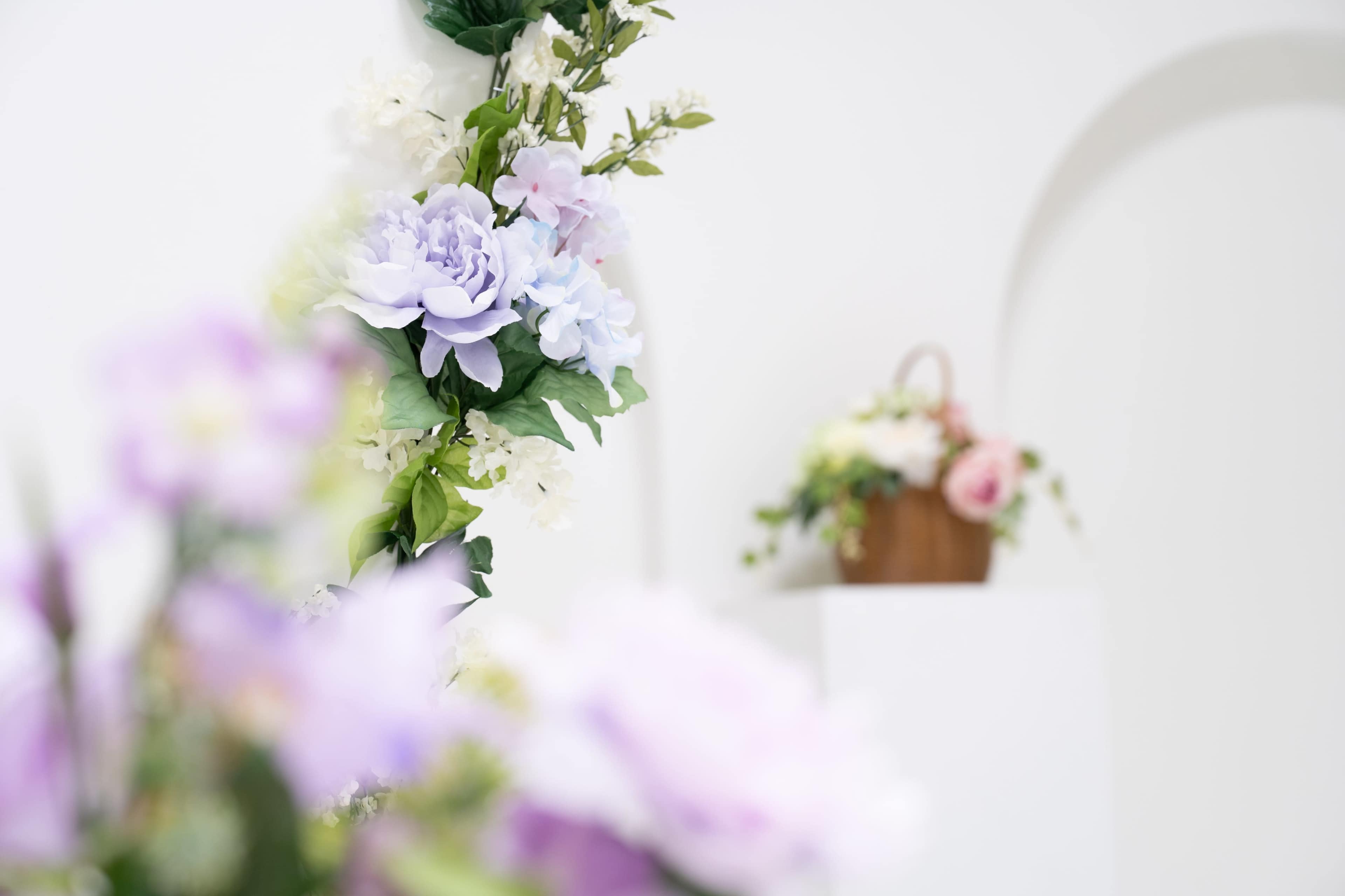 A cascade of artificial flowers in shades of purple and white decorates a white wall, with a decorative basket of blooms positioned on a pedestal in the background.