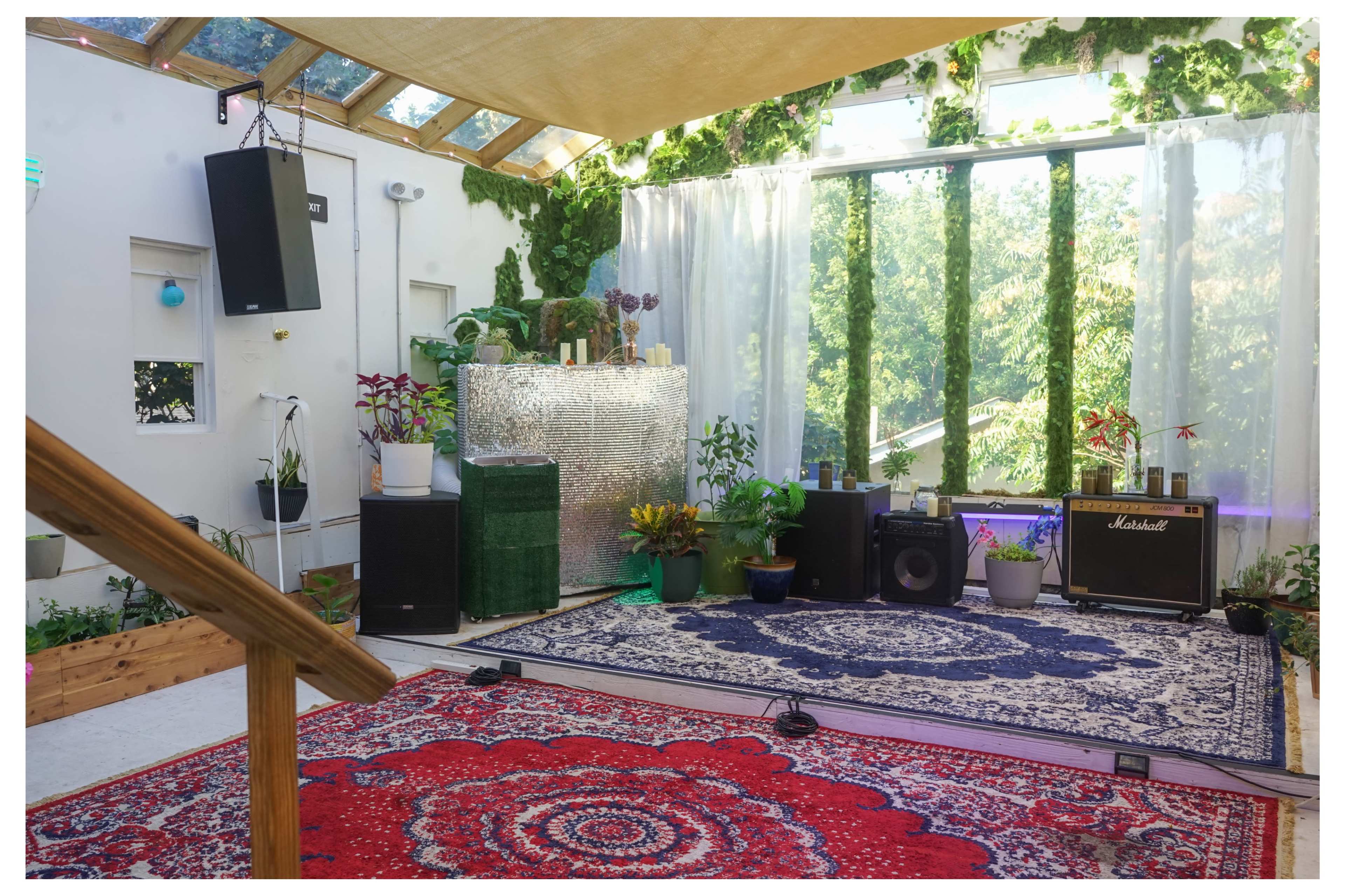 The interior of a room features large windows, various plants, and audio equipment on a patterned rug.