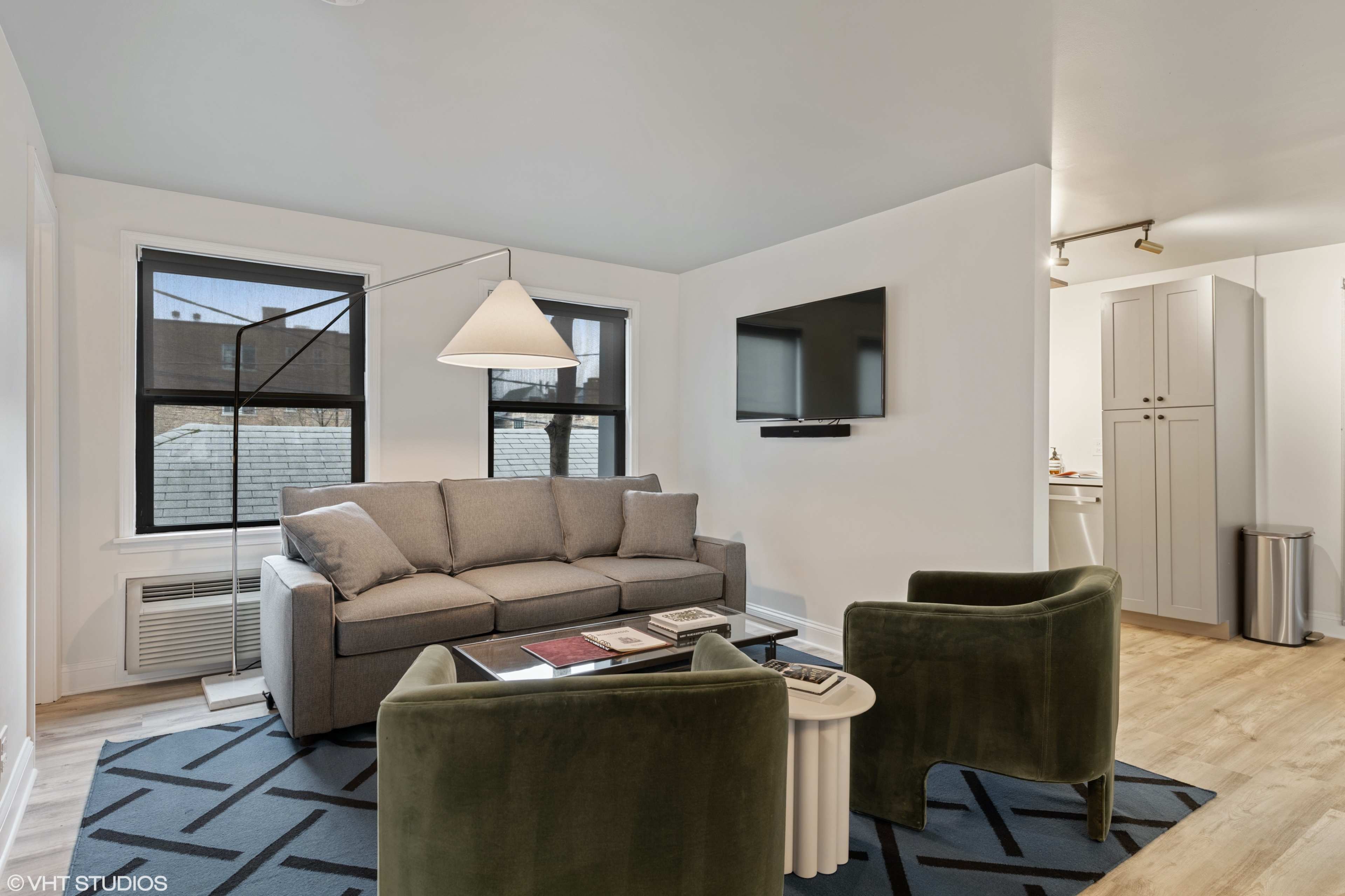 A modern living room with a gray sofa, two green armchairs, a coffee table, and a wall-mounted television, with a kitchenette visible in the background.