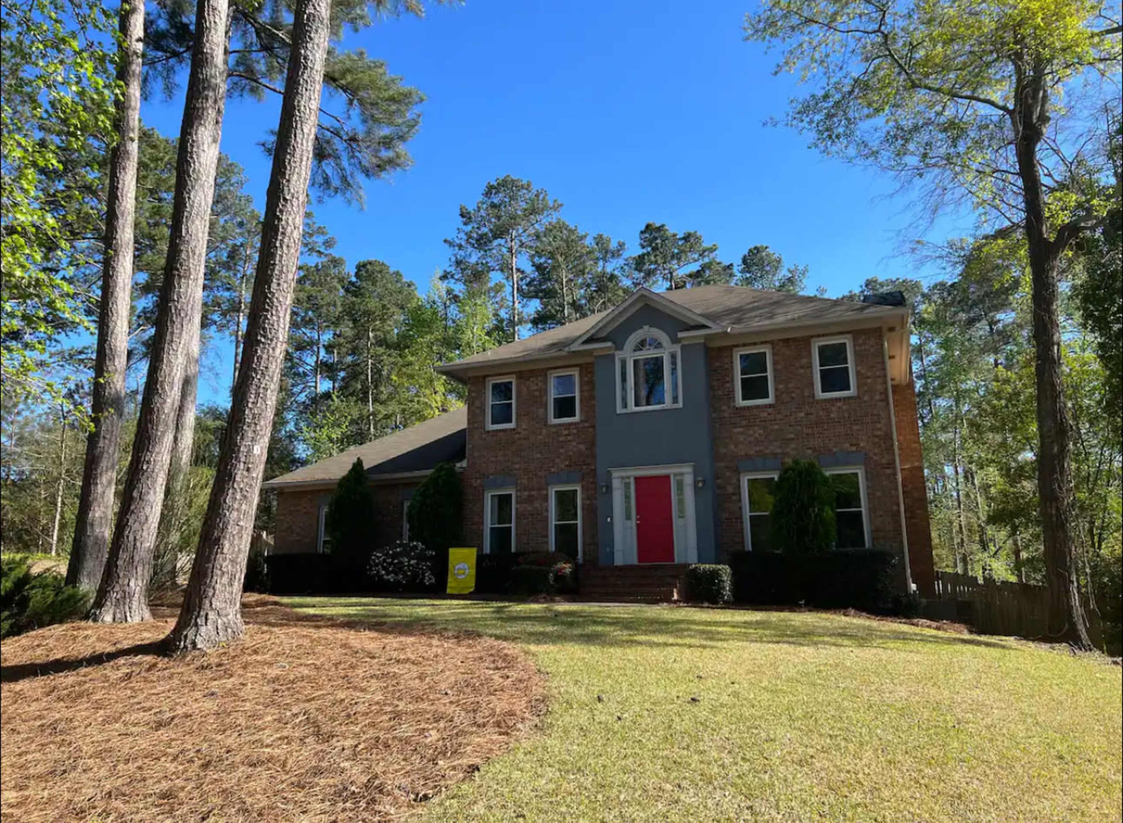 The image shows a two-story brick house with a red front door, surrounded by pine trees and a manicured lawn.