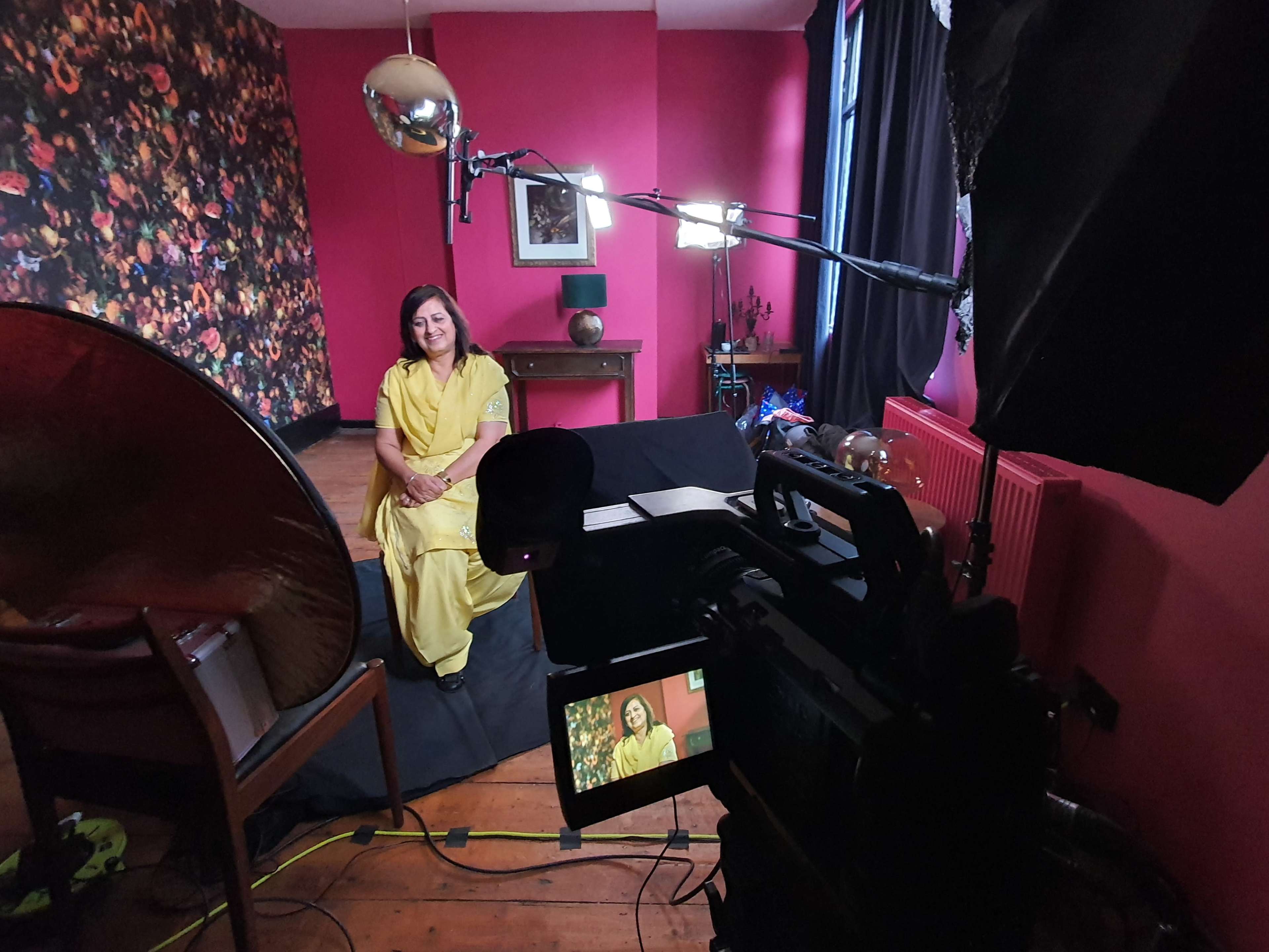 A woman in a yellow outfit sits on a black couch in a room with pink walls and floral wallpaper, while a camera and lighting equipment are set up for recording.