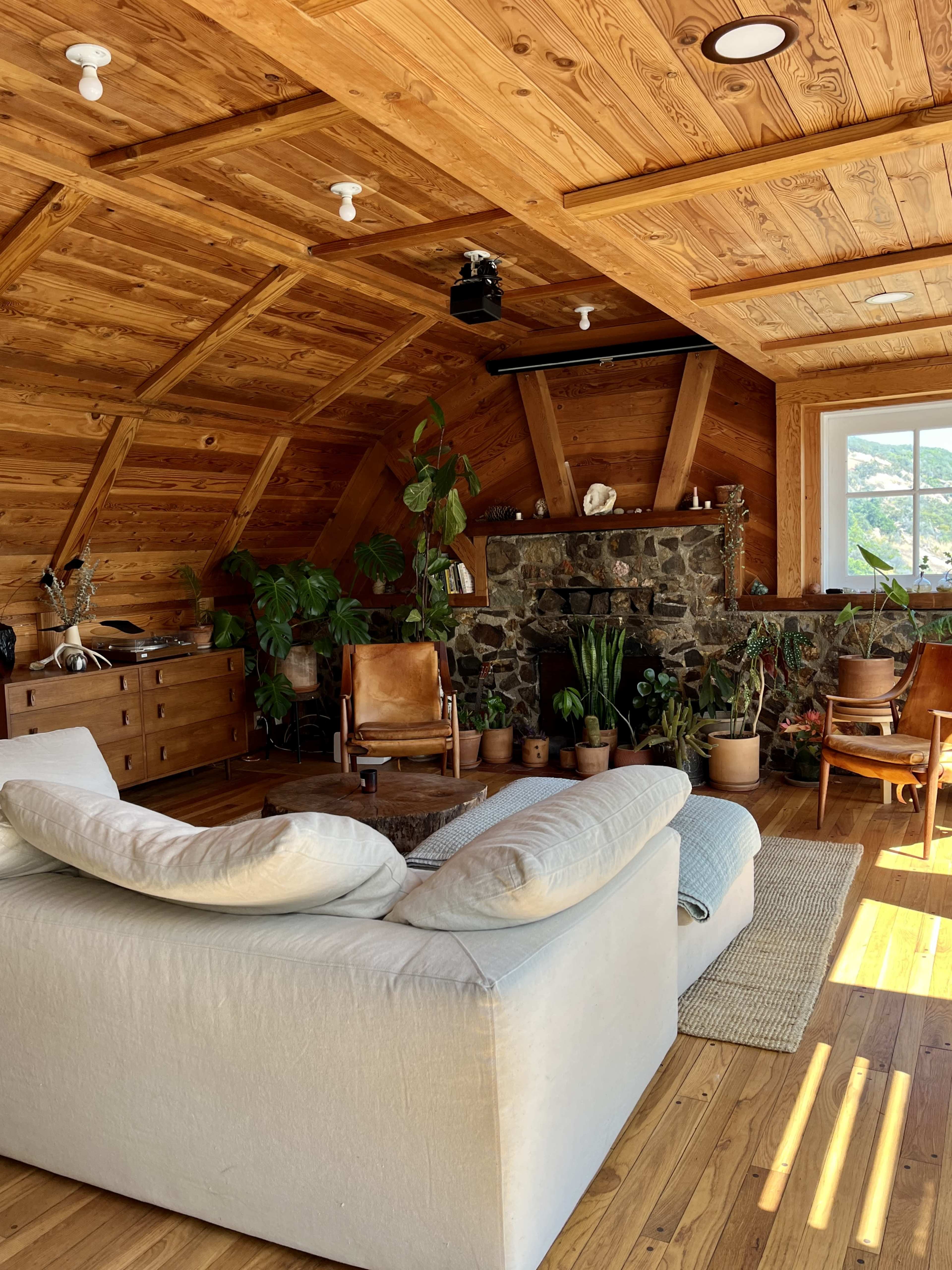 A cozy wooden interior with a stone fireplace, a white sofa, several potted plants, and natural light streaming through a large window.