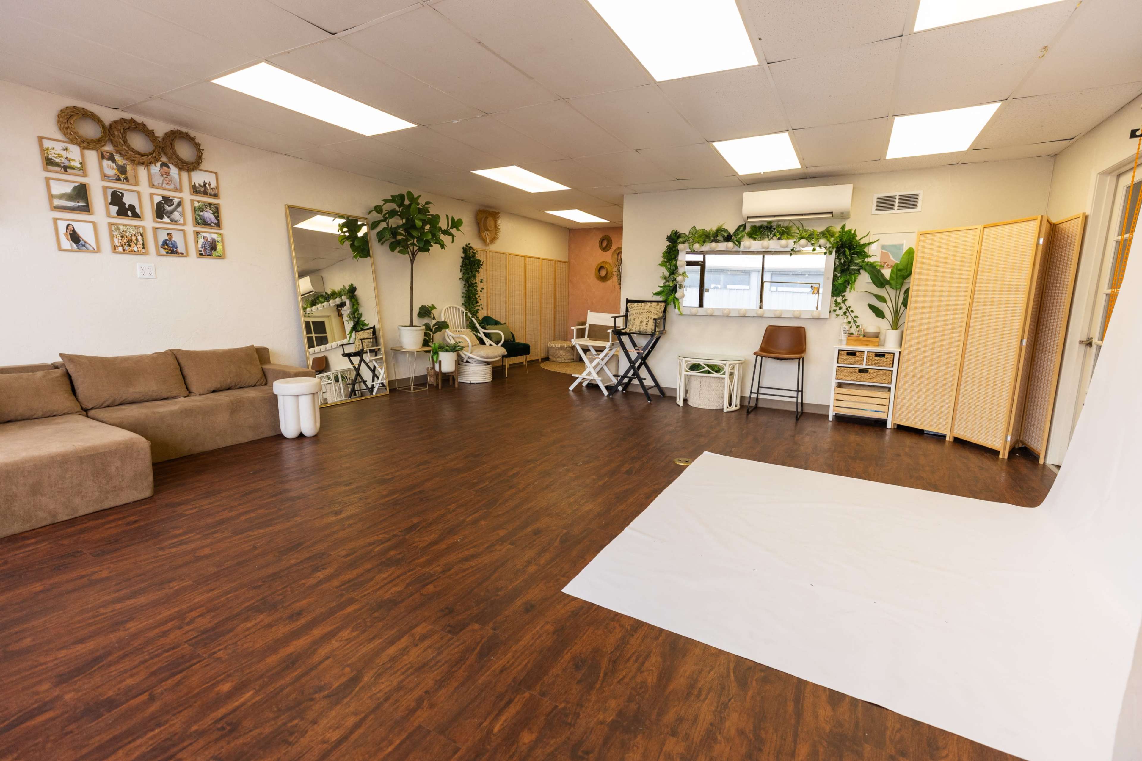 The image shows a spacious studio with wooden flooring, a large white backdrop, seating arrangements, plants, and decorative wall art.
