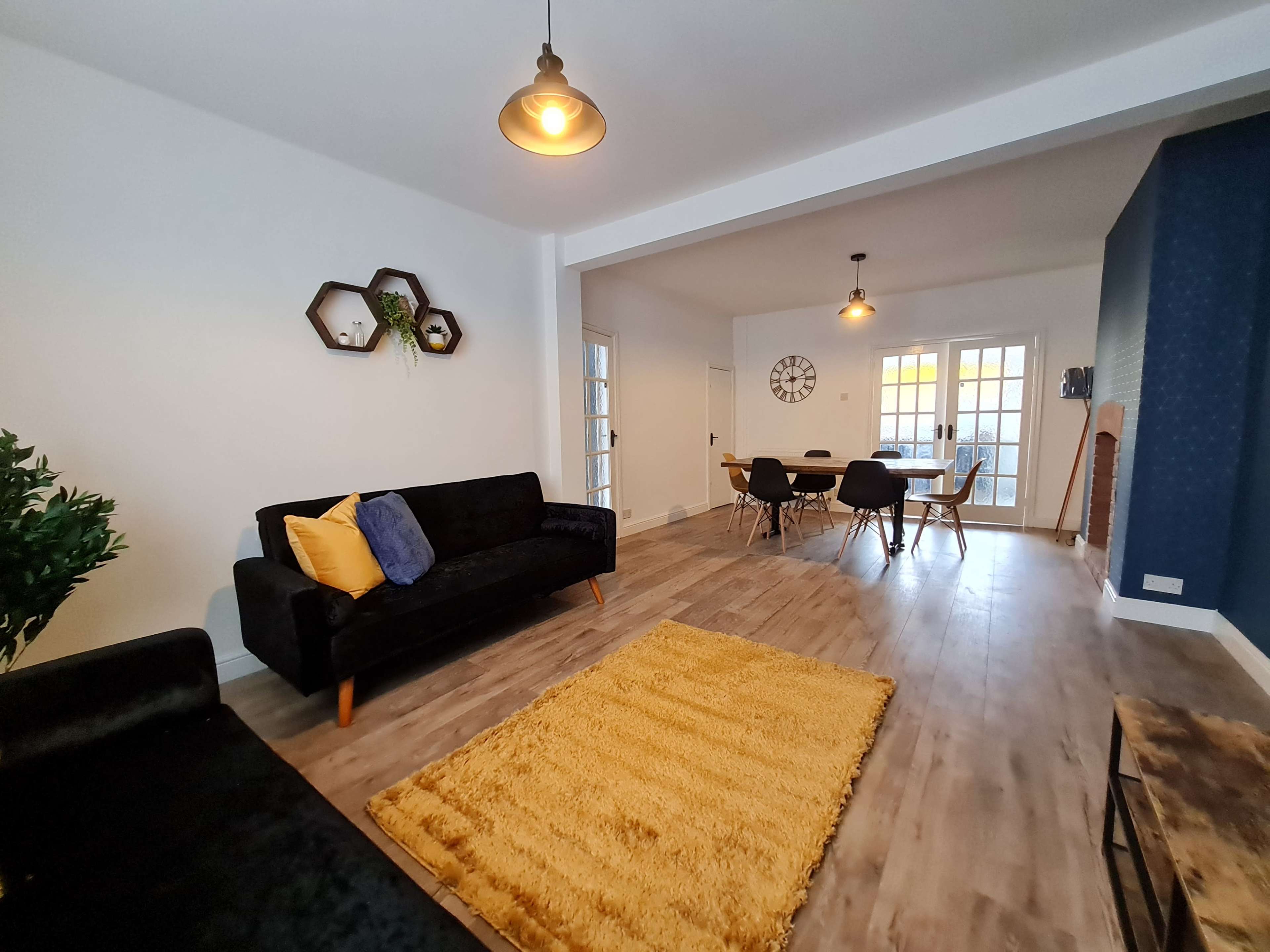 A modern living room features a black sofa, a yellow rug, and a dining area with a wooden table and chairs, illuminated by pendant lights.
