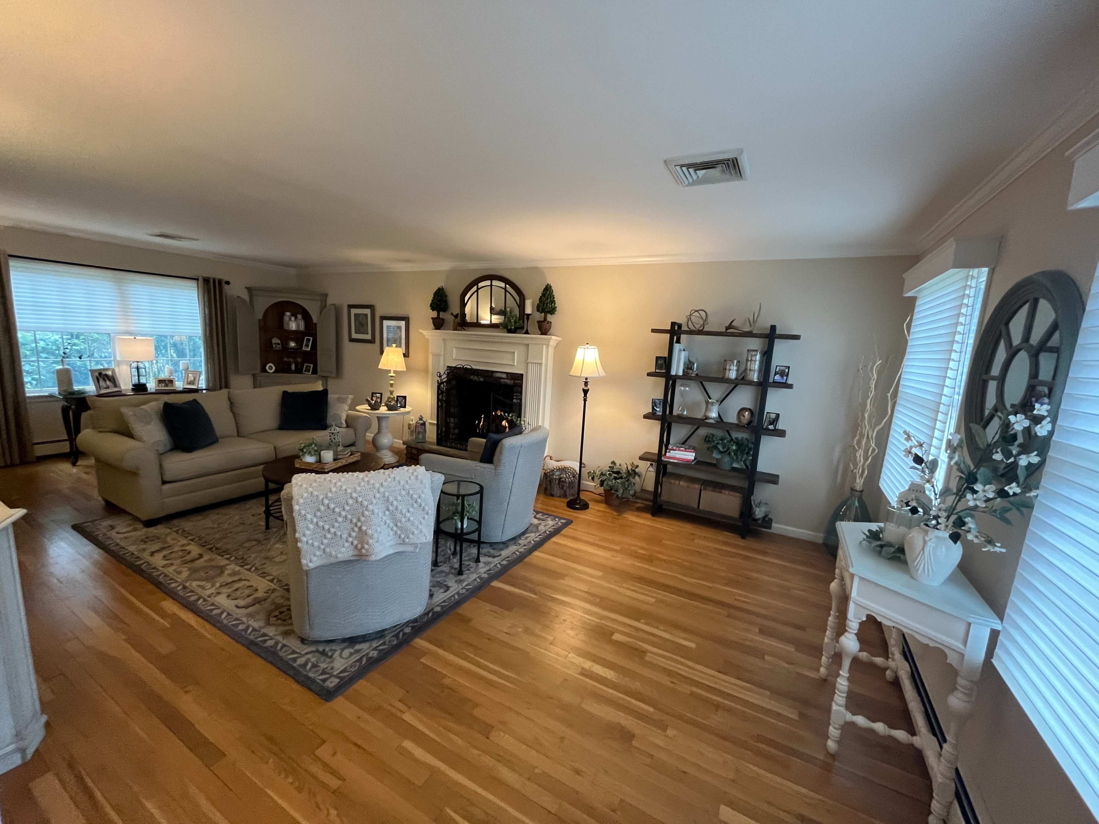 A spacious living room features a sofa, a coffee table, a fireplace, shelves with decor, and large windows allowing natural light.