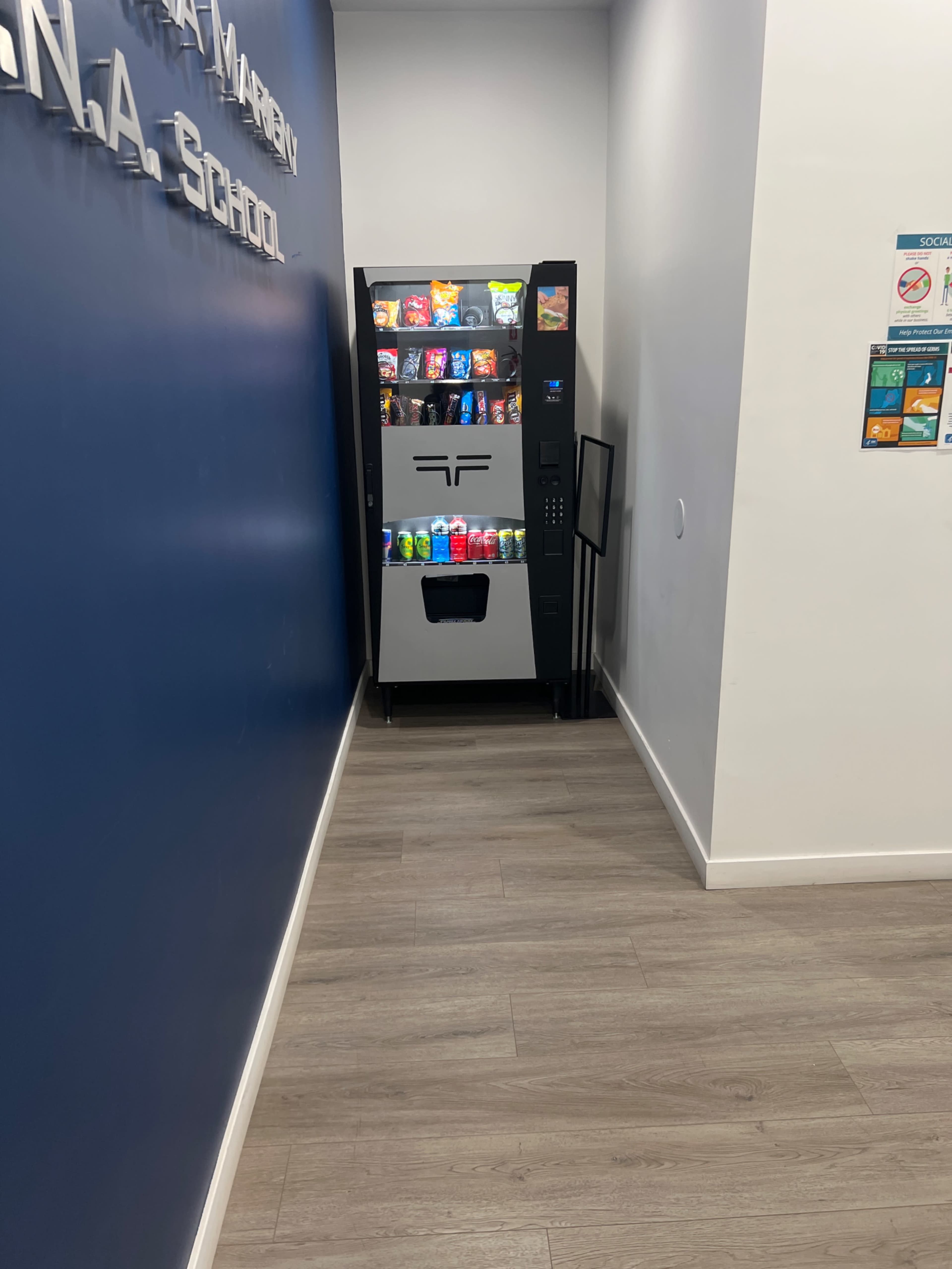 A vending machine filled with snacks and beverages is located in a corridor beside a blue wall.