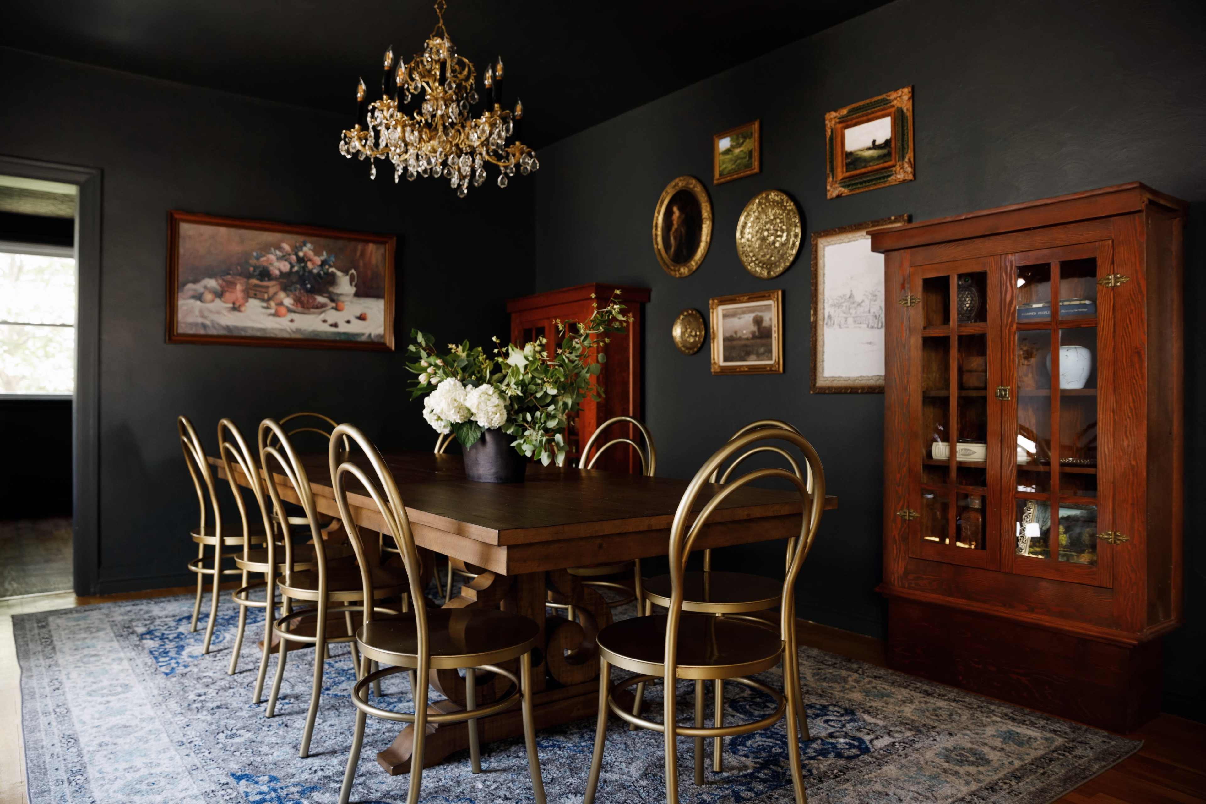 A spacious dining room features a large wooden table surrounded by gold chairs, with a crystal chandelier overhead and framed artwork on the dark walls.