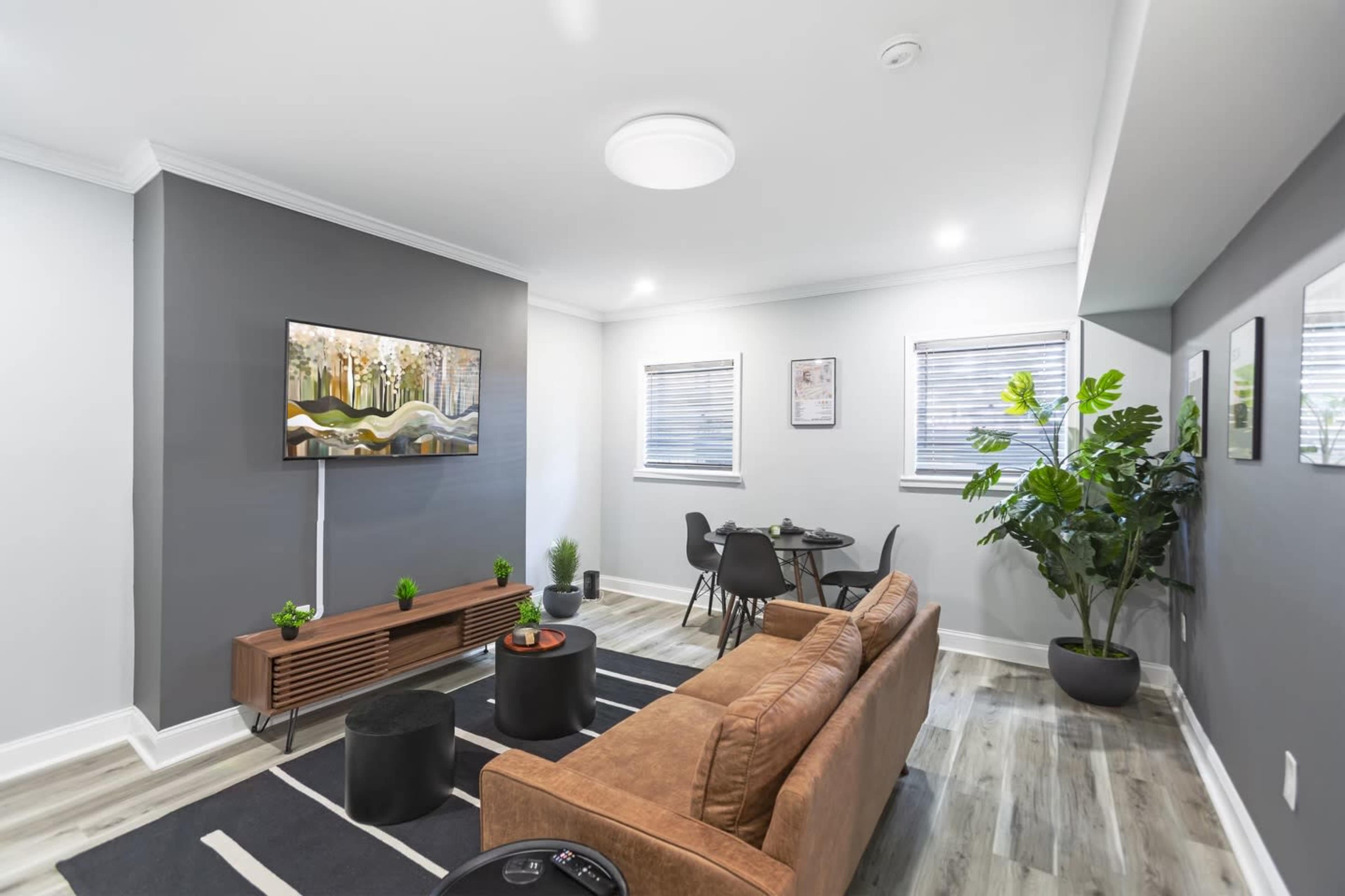 A modern living room features a brown sofa, a television mounted on the wall, a small dining table, and a large potted plant.