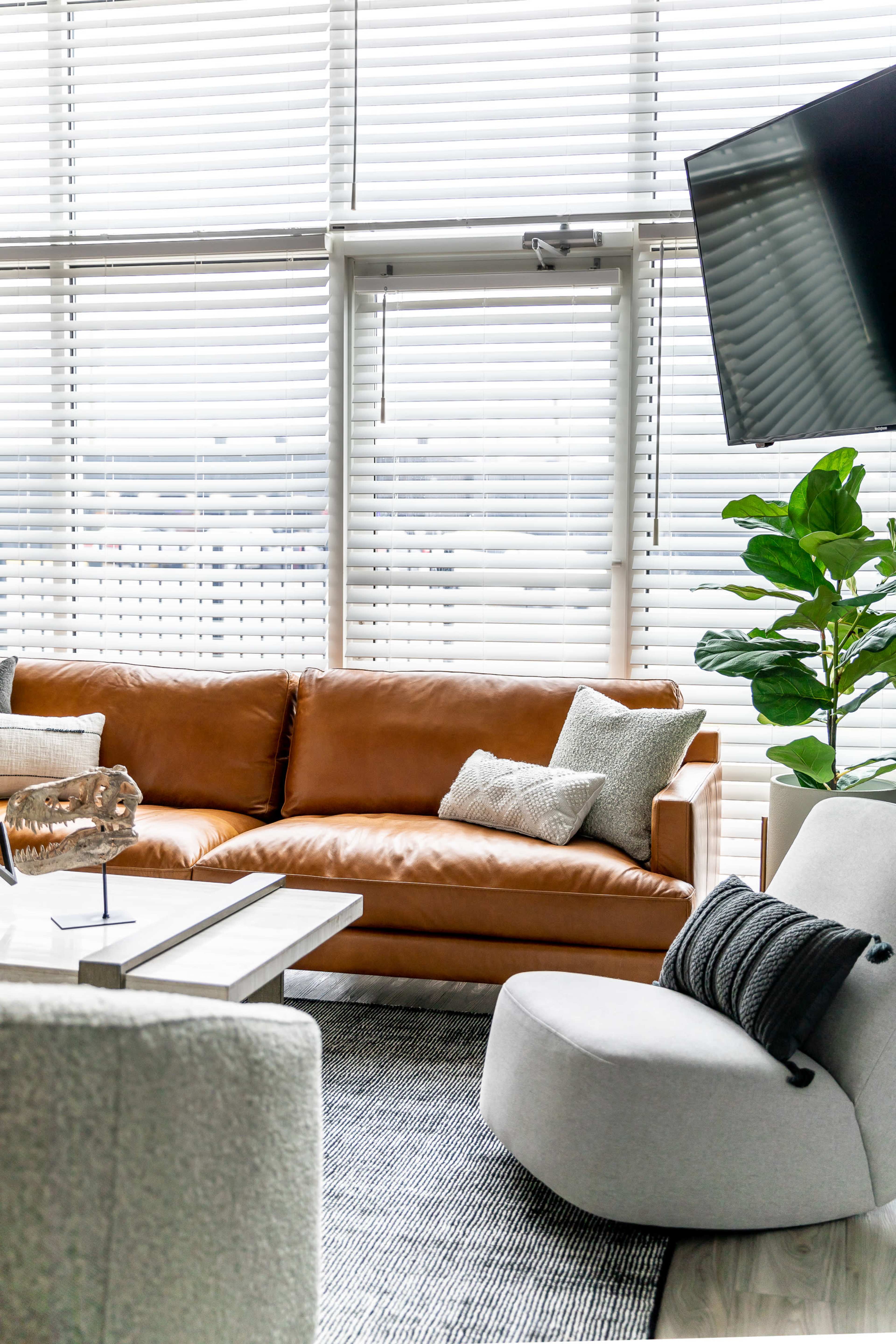 A modern living room featuring a brown leather sofa, a gray accent chair, a coffee table, and large windows with blinds.