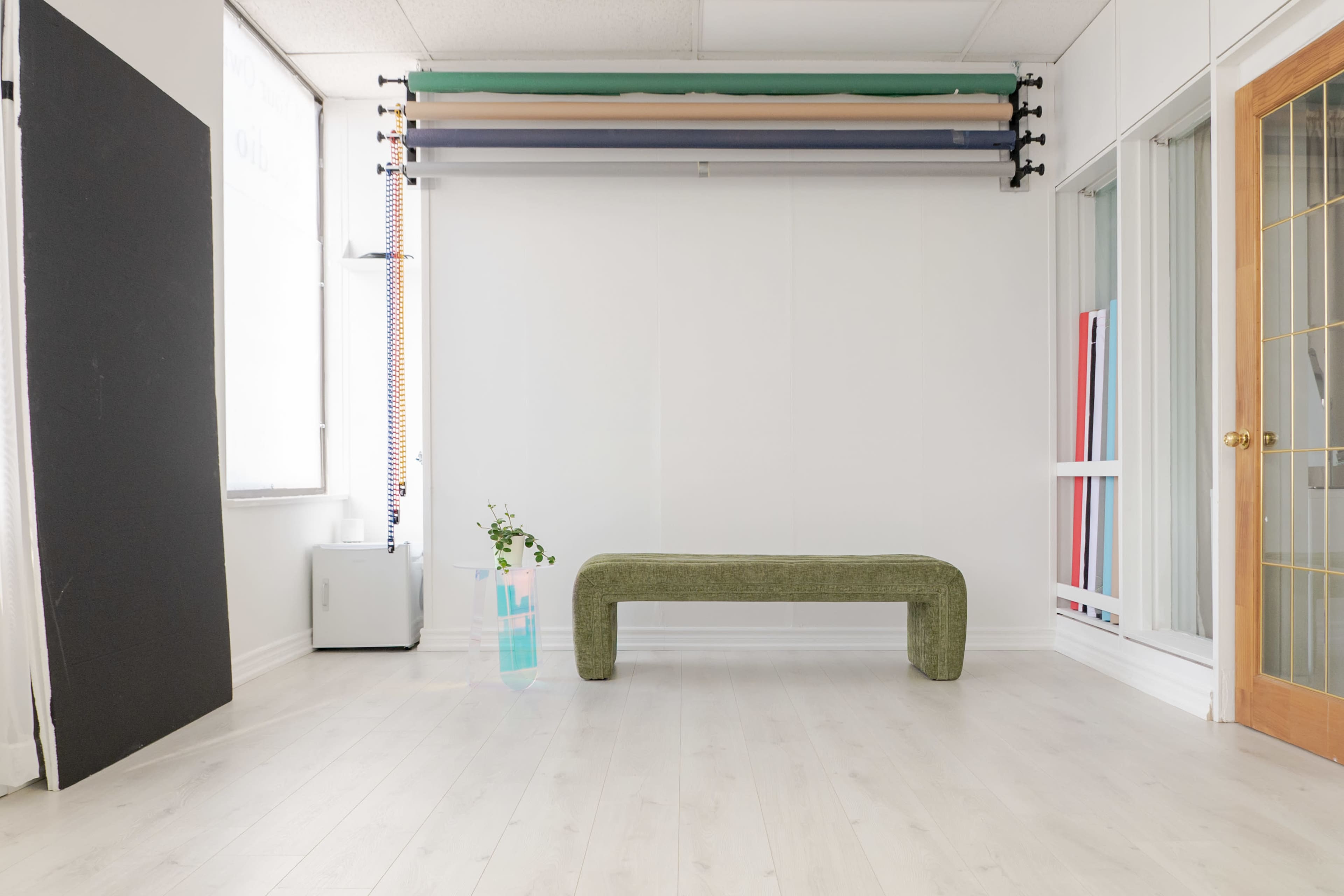 The image shows a minimalist studio space with a green bench, a small plant, and backdrop rolls mounted on the wall.