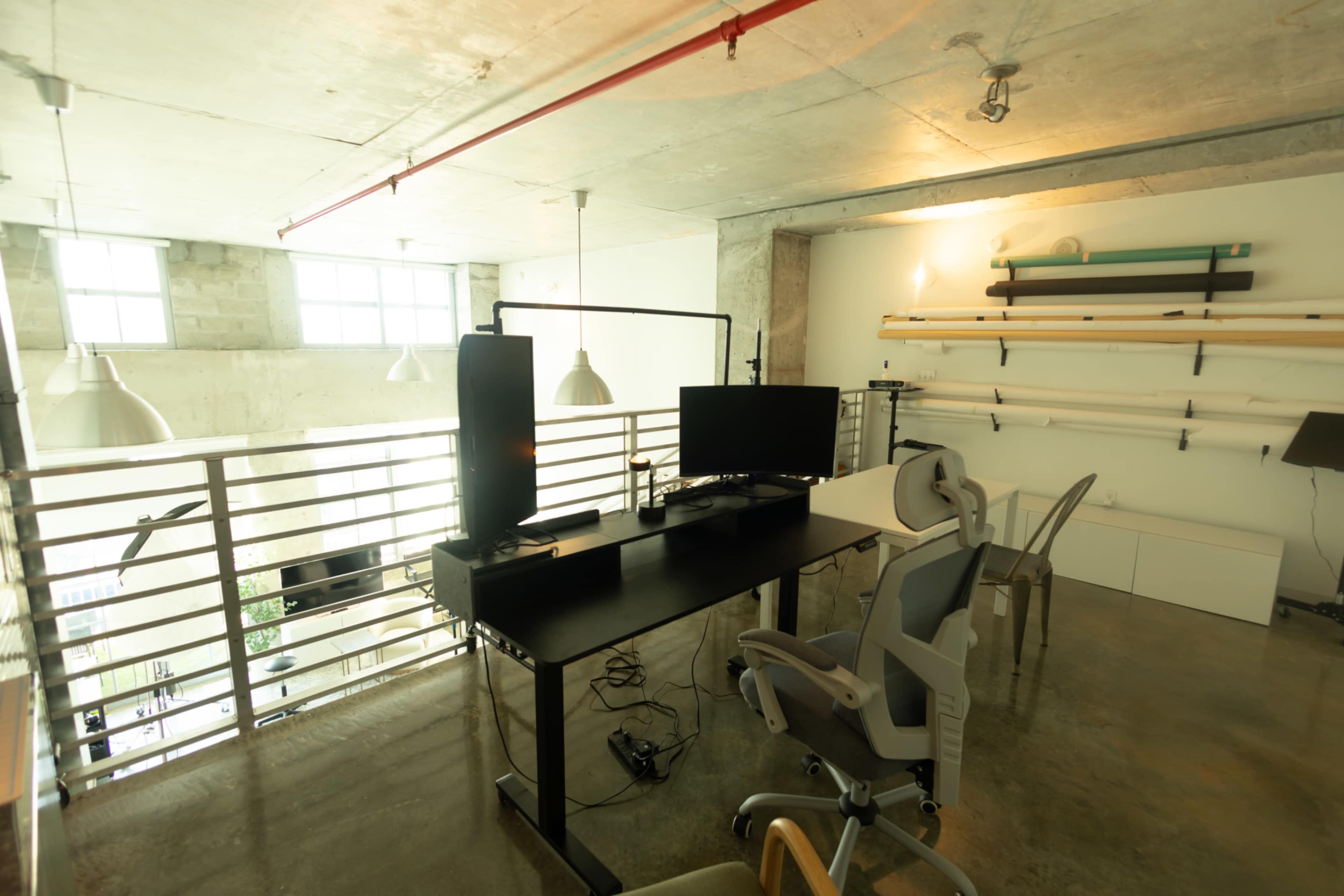 The image shows a modern office space featuring a desk with a computer monitor, an ergonomic chair, and exposed concrete walls, along with large windows allowing natural light to enter.