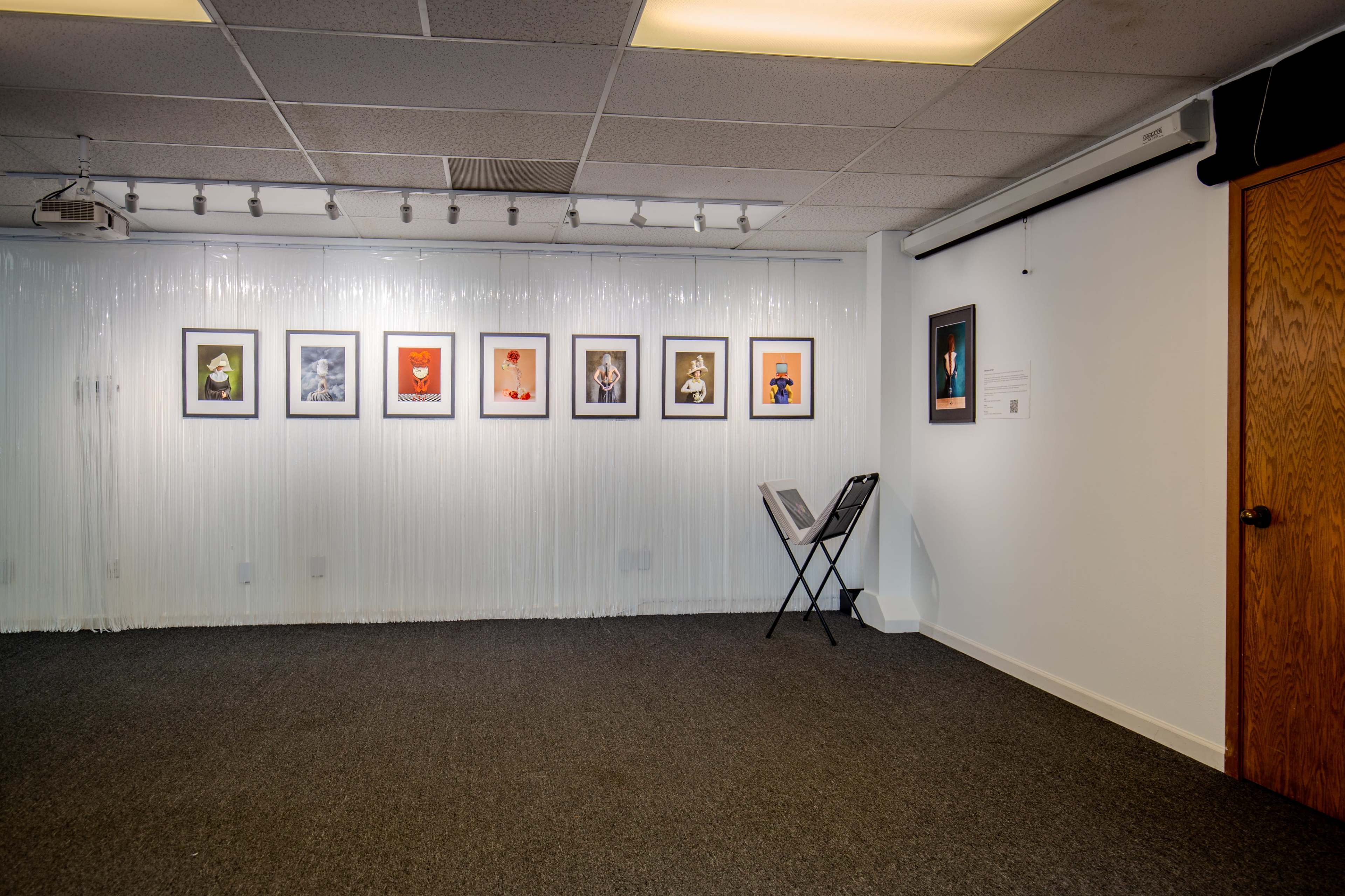 The image shows a gallery room with a series of framed artworks hanging on a white wall, a chair positioned on the carpeted floor, and a partially opaque curtain in the background.