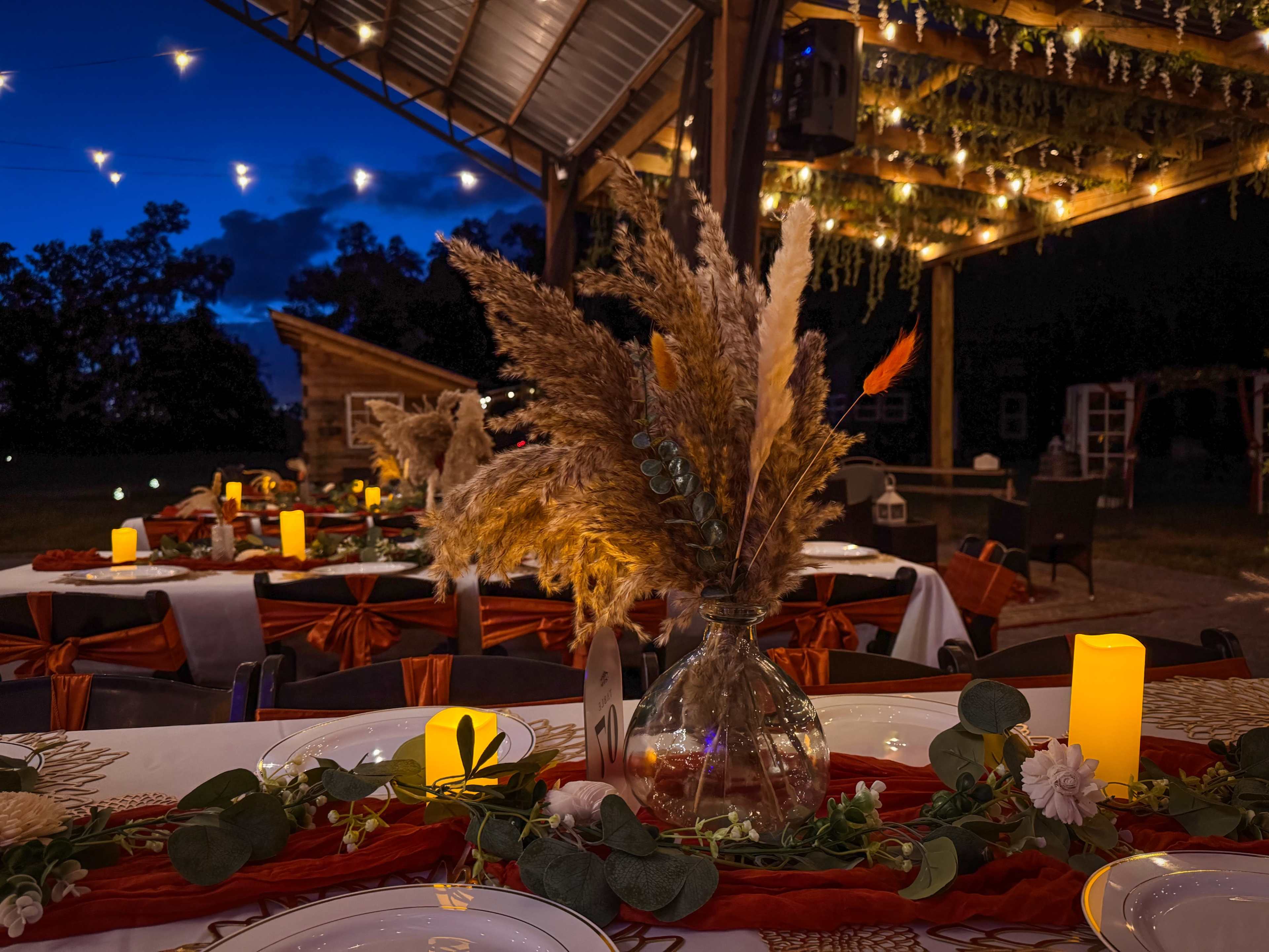 The image shows a beautifully arranged outdoor dining setup under a canopy with warm lighting, featuring decorated tables with floral centerpieces.
