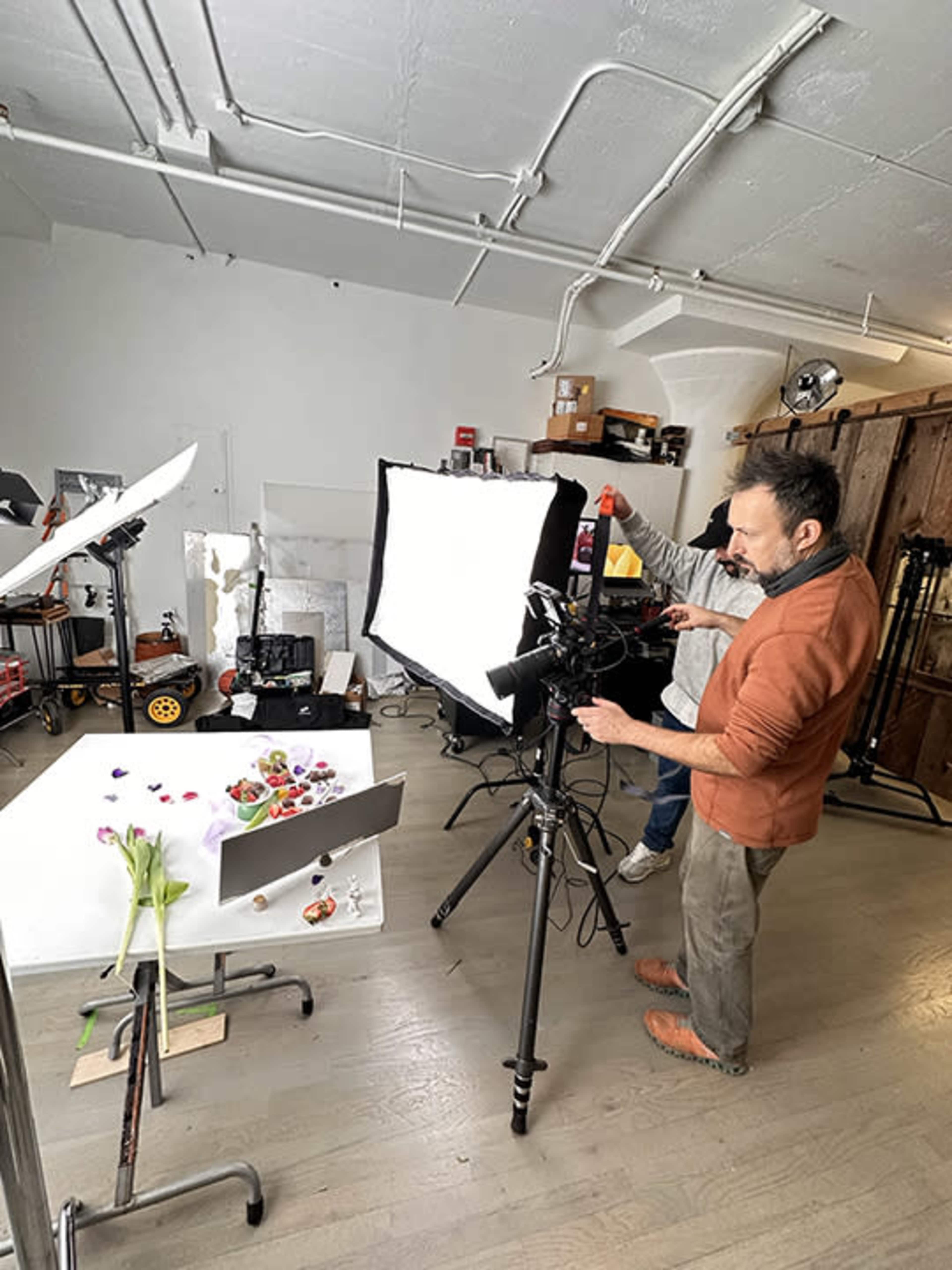 A photographer adjusts equipment in a studio filled with lighting setups and a variety of colorful objects on a table.