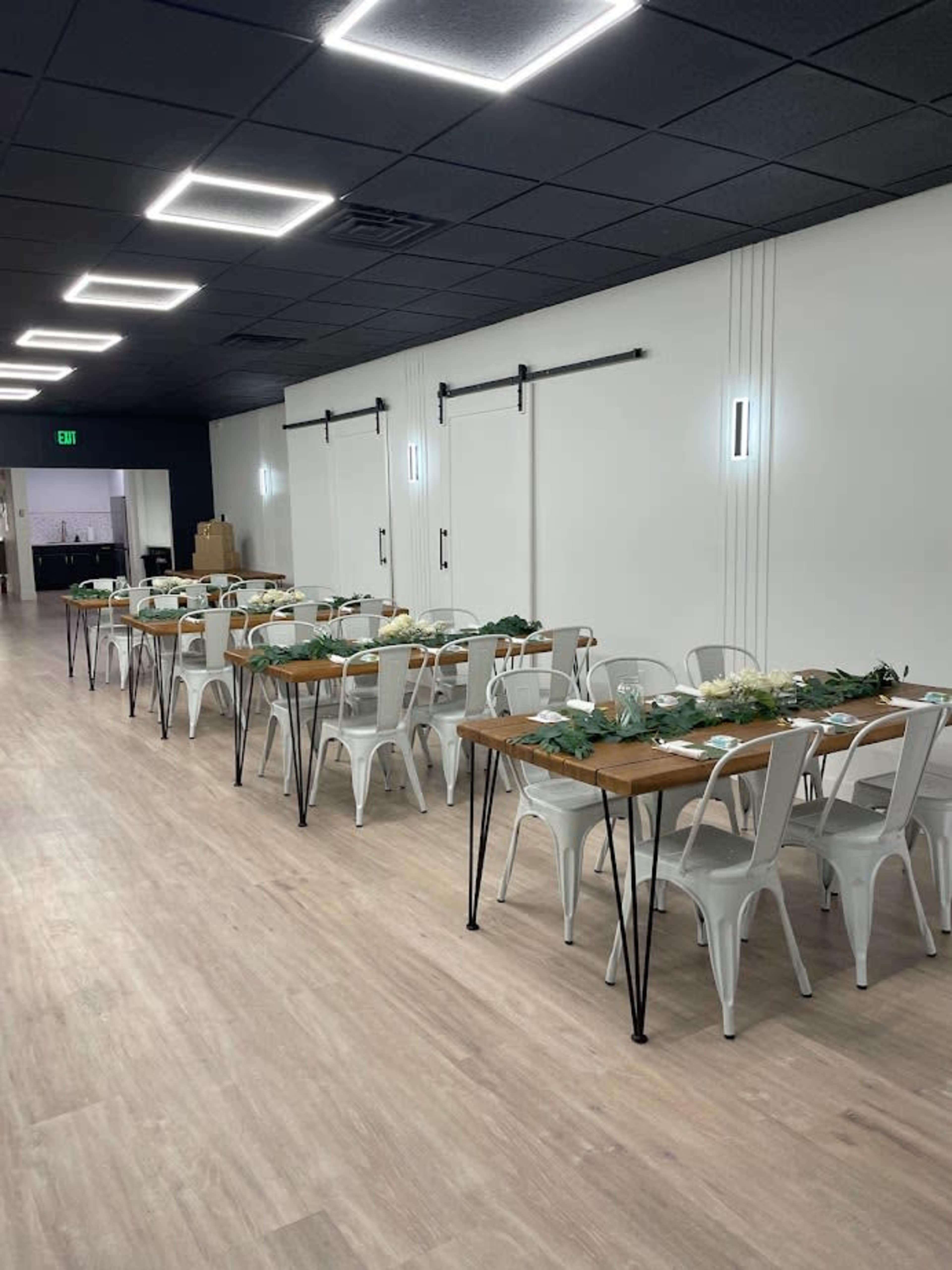 A long, rectangular dining setup with multiple tables lined with greenery and flowers is arranged in a well-lit room featuring white walls and modern lighting.