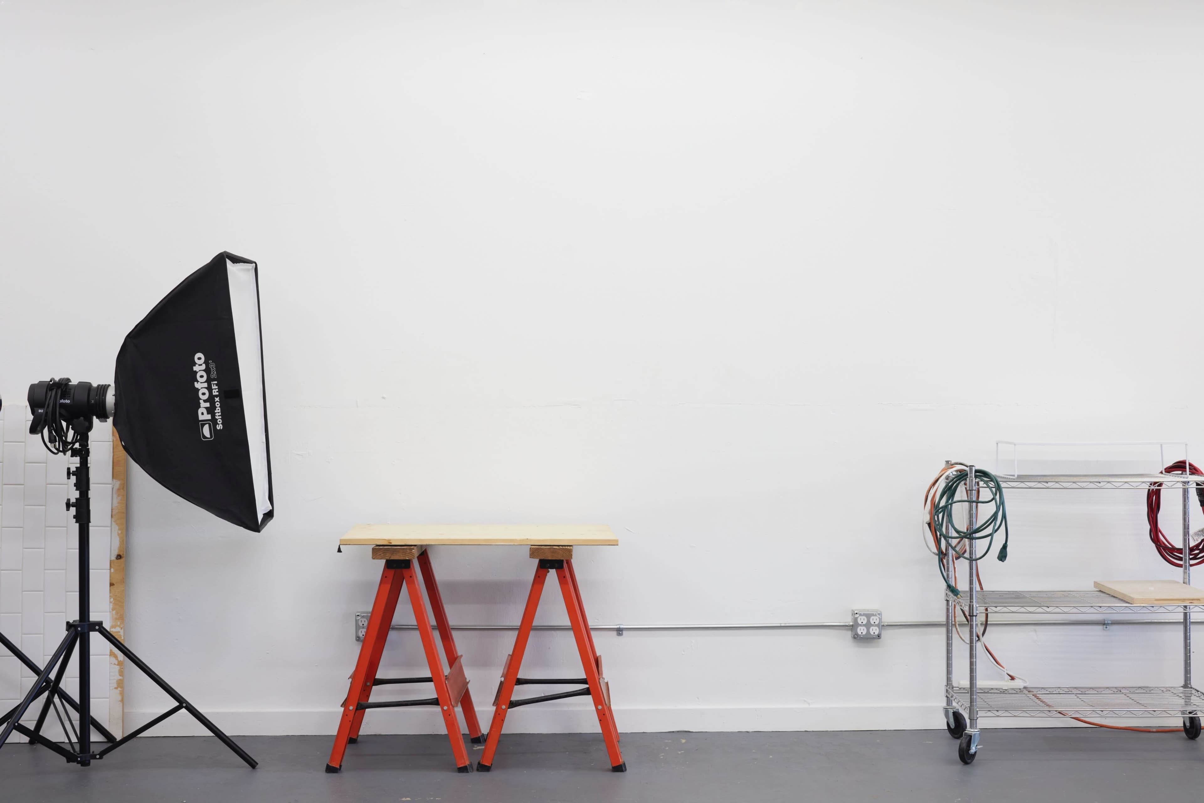 A minimalist studio setup with a light source on a tripod, a wooden work table, and a metal shelving unit with cables beside a plain white wall.