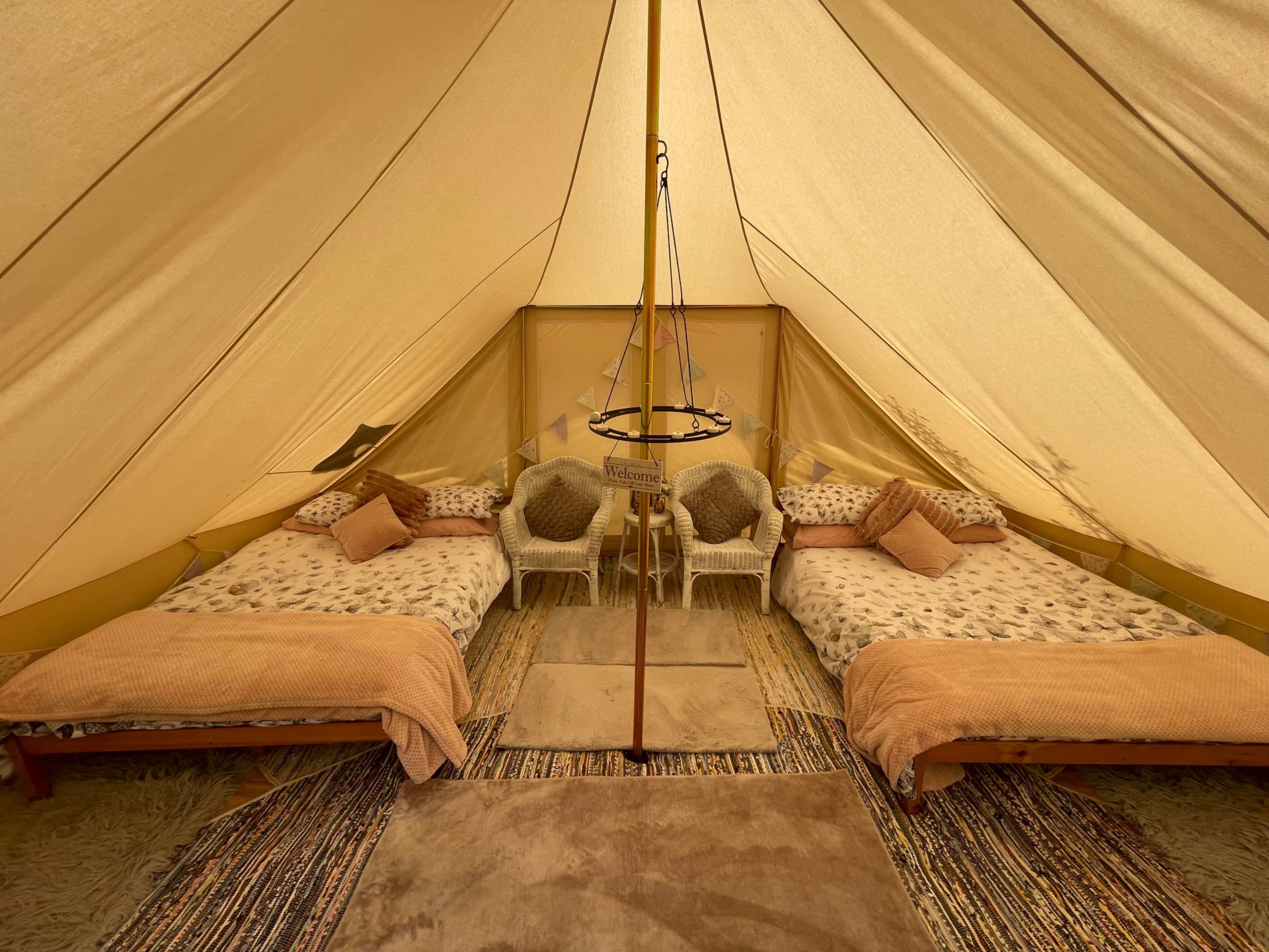 A spacious, tent interior with two made beds, decorative pillows, and a central hanging light fixture.