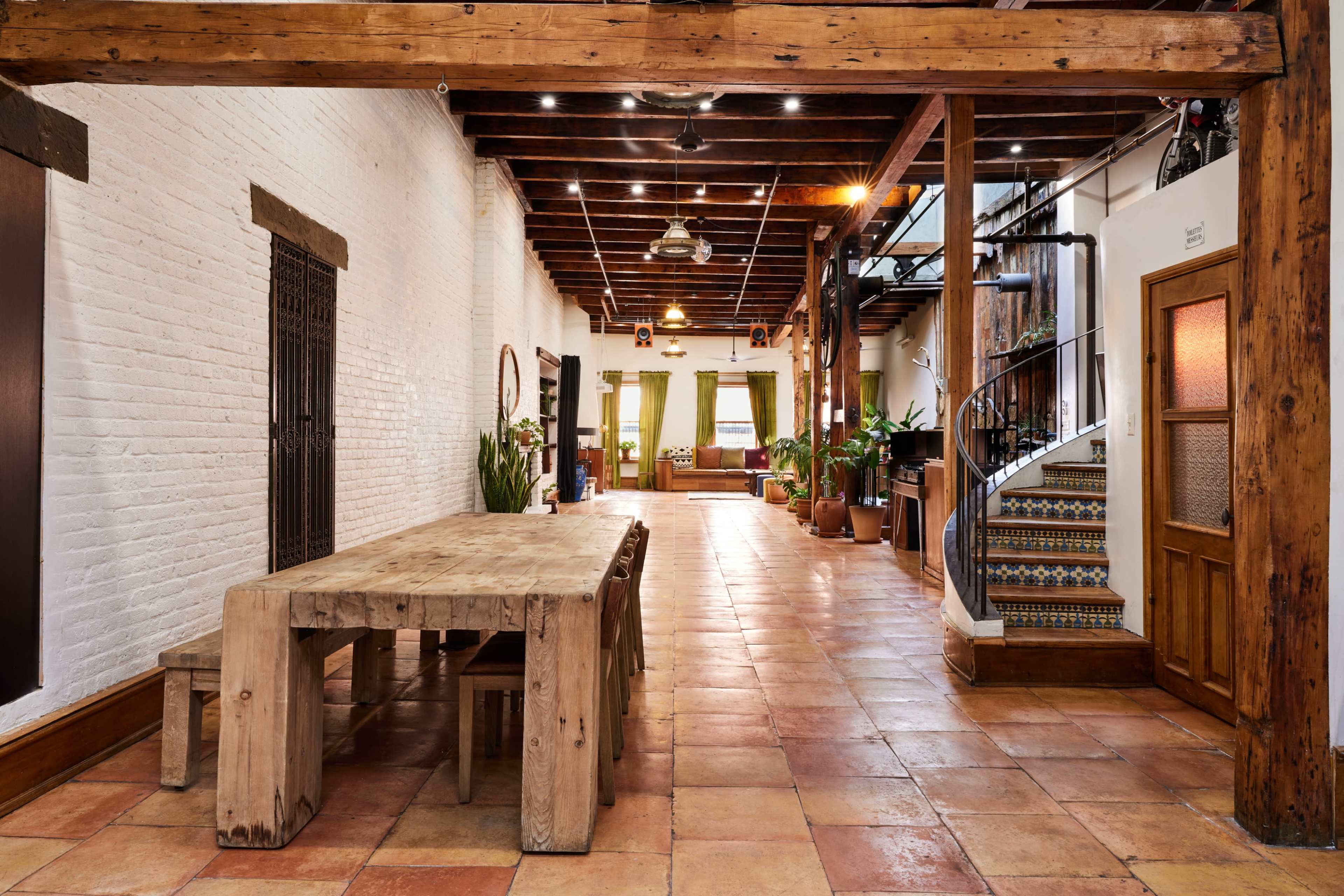 A long, narrow interior space with a wooden dining table, exposed wooden beams, and decorative tile stairs, leading to a living area with plants and large windows.