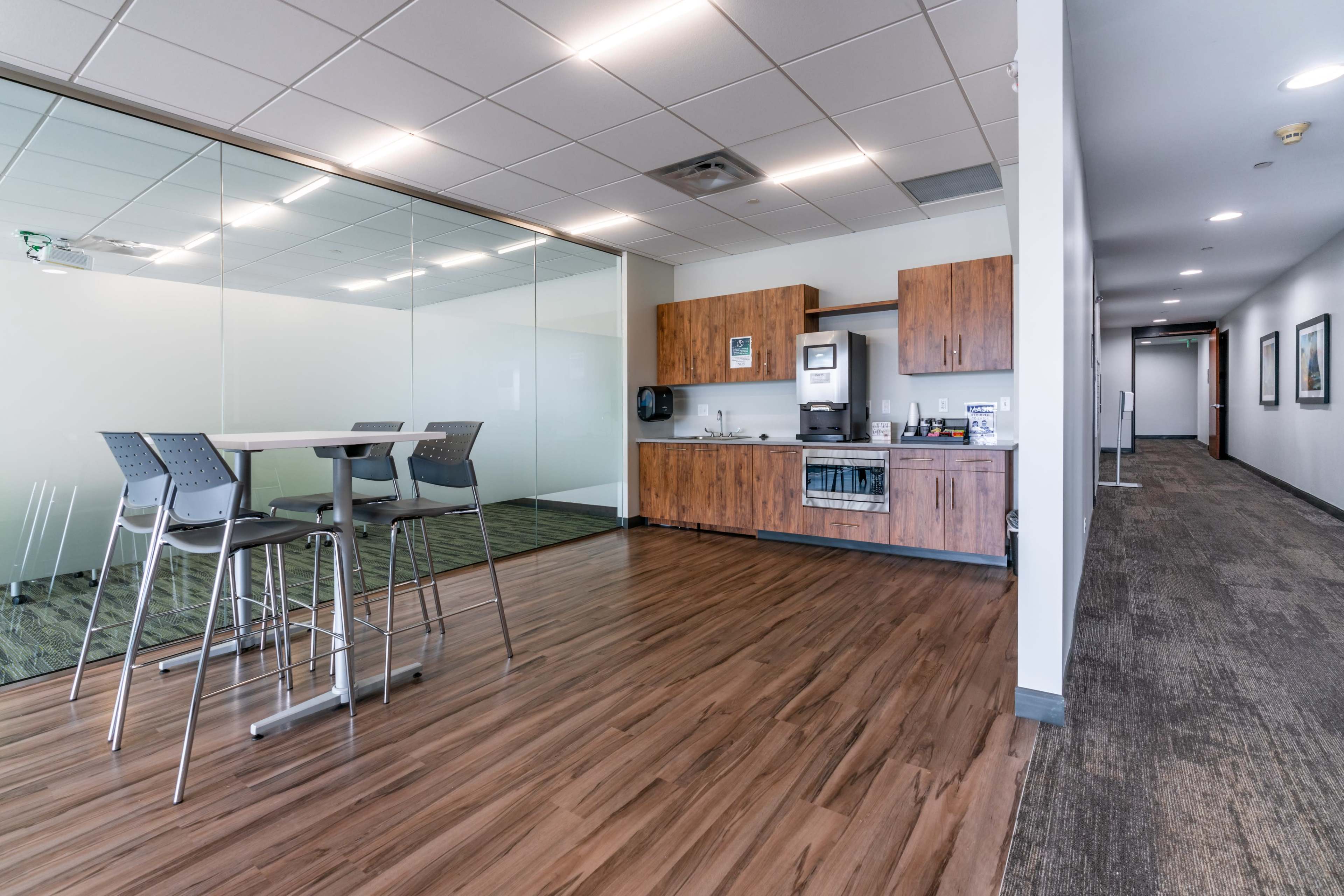 A modern office break area with a kitchenette, tall table and chairs, and a hallway leading to closed office spaces.