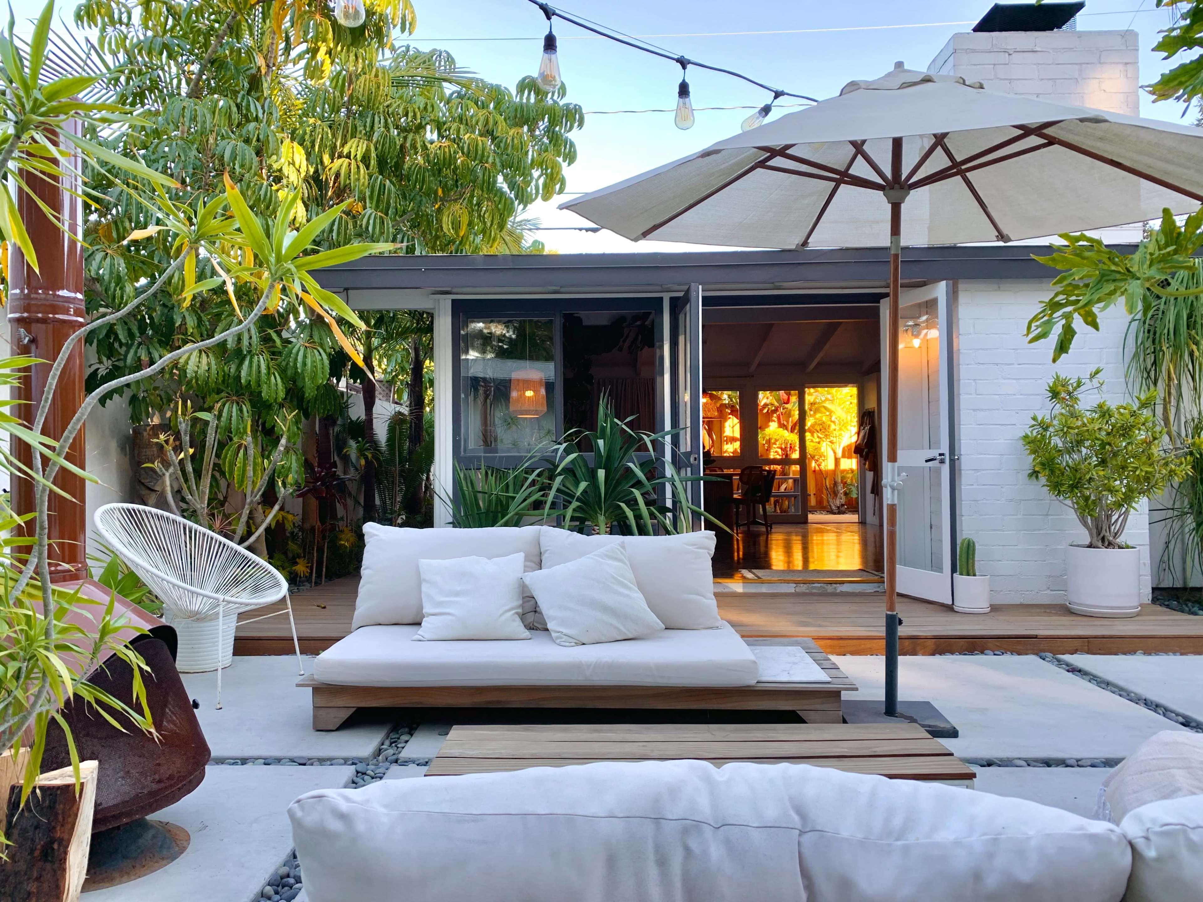 A cozy outdoor seating area with a large umbrella, surrounded by plants, leads to a well-lit interior space through glass doors.