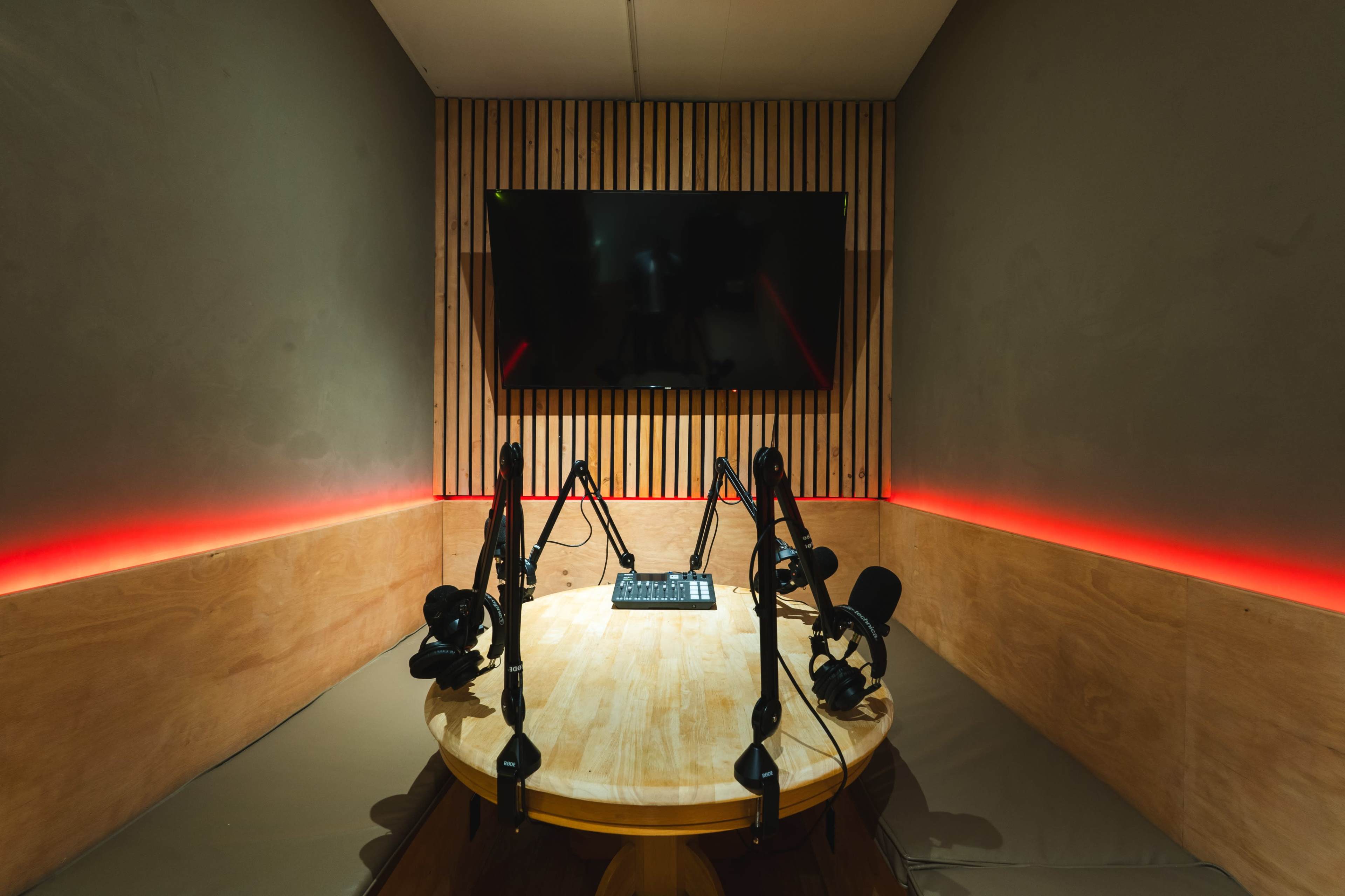 A recording studio with a round wooden table, multiple microphones, a control panel, and a large flat-screen TV mounted on a wooden paneled wall, illuminated by red LED lighting.