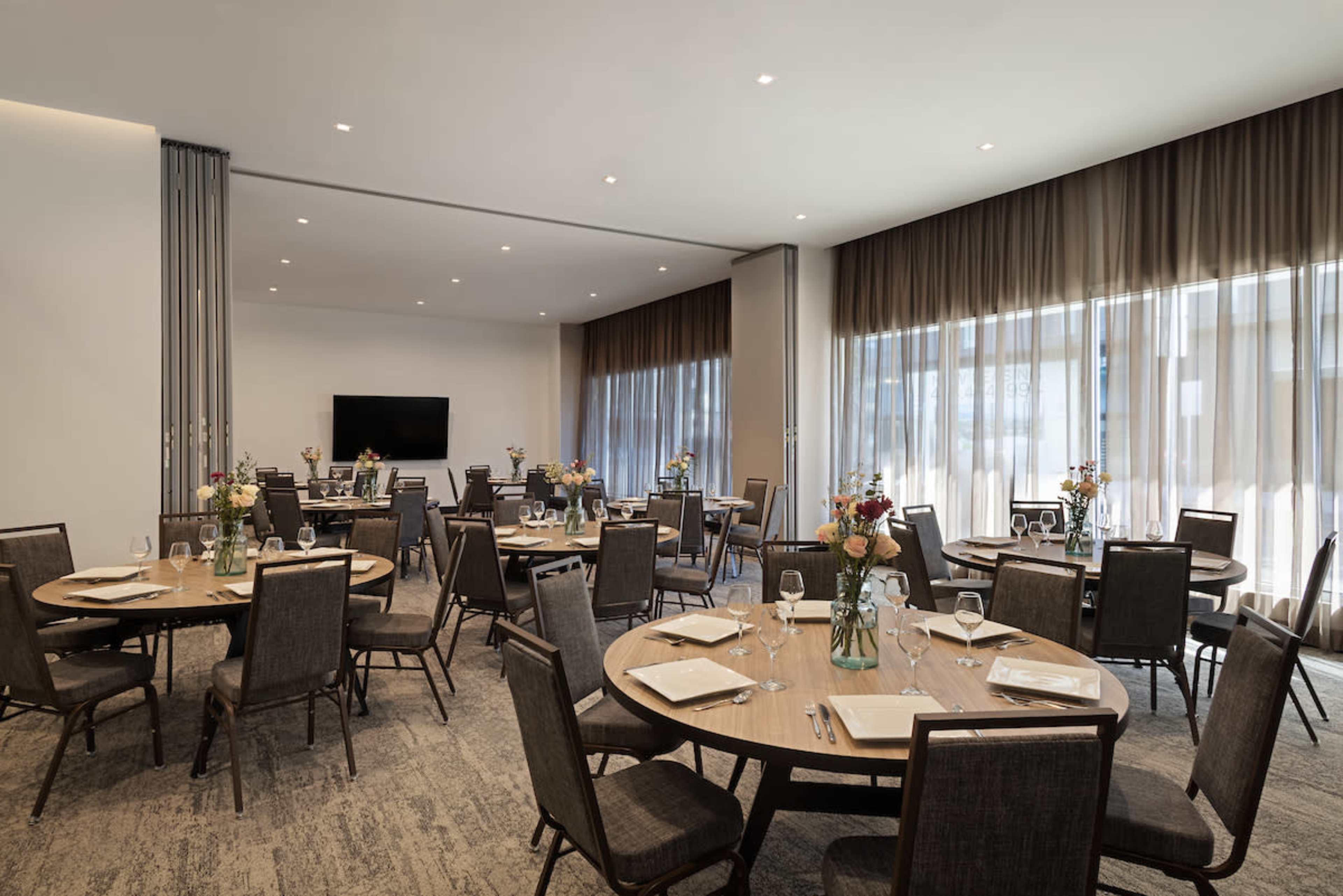 The image shows a spacious dining room set with round tables arranged neatly, each adorned with flower centerpieces and table settings.