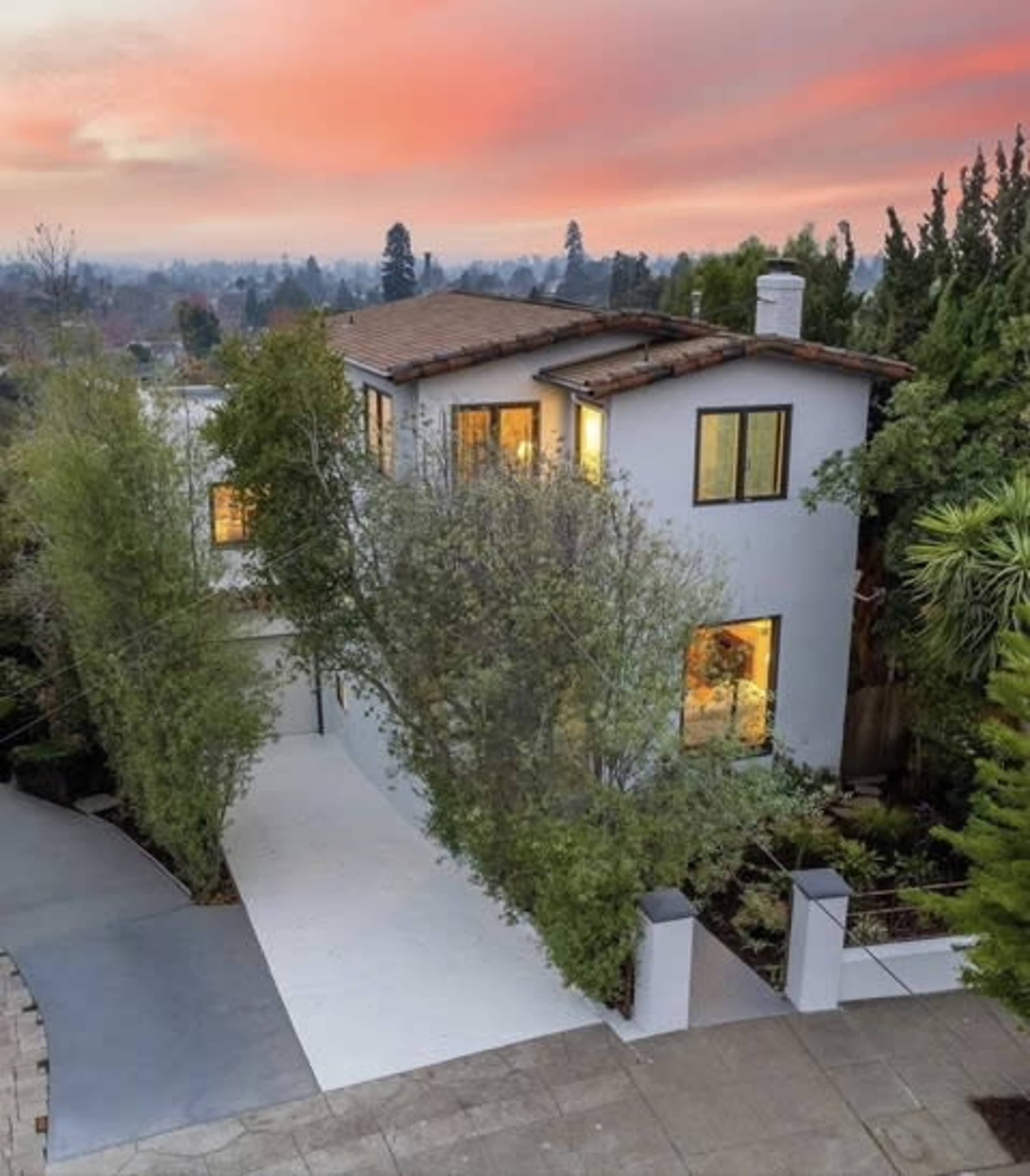 A modern two-story house with a sloped roof is surrounded by greenery, featuring large windows that reflect a colorful sunset sky.