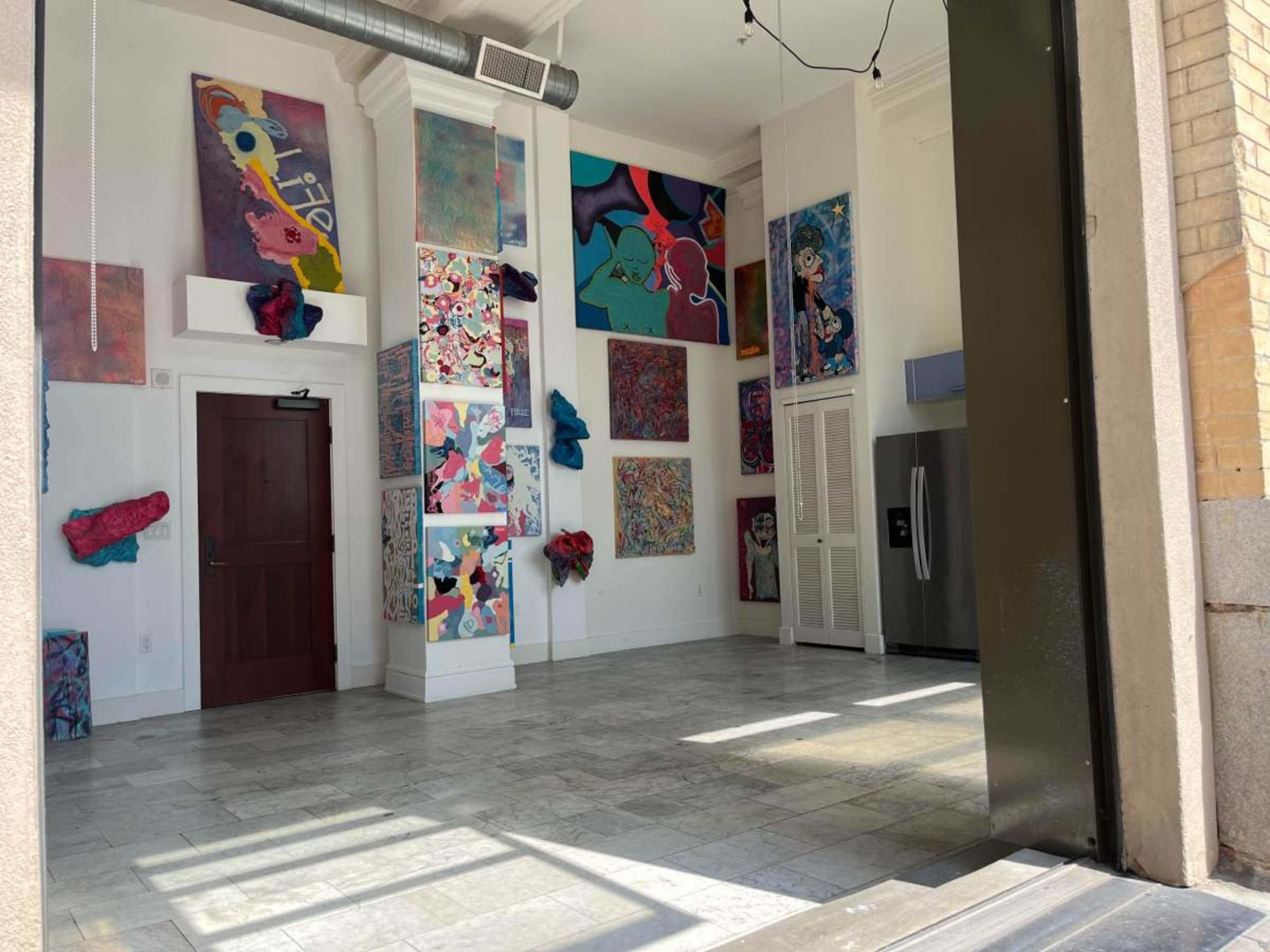 The image shows an empty room with colorful abstract artwork displayed on the walls and a refrigerator in the corner.